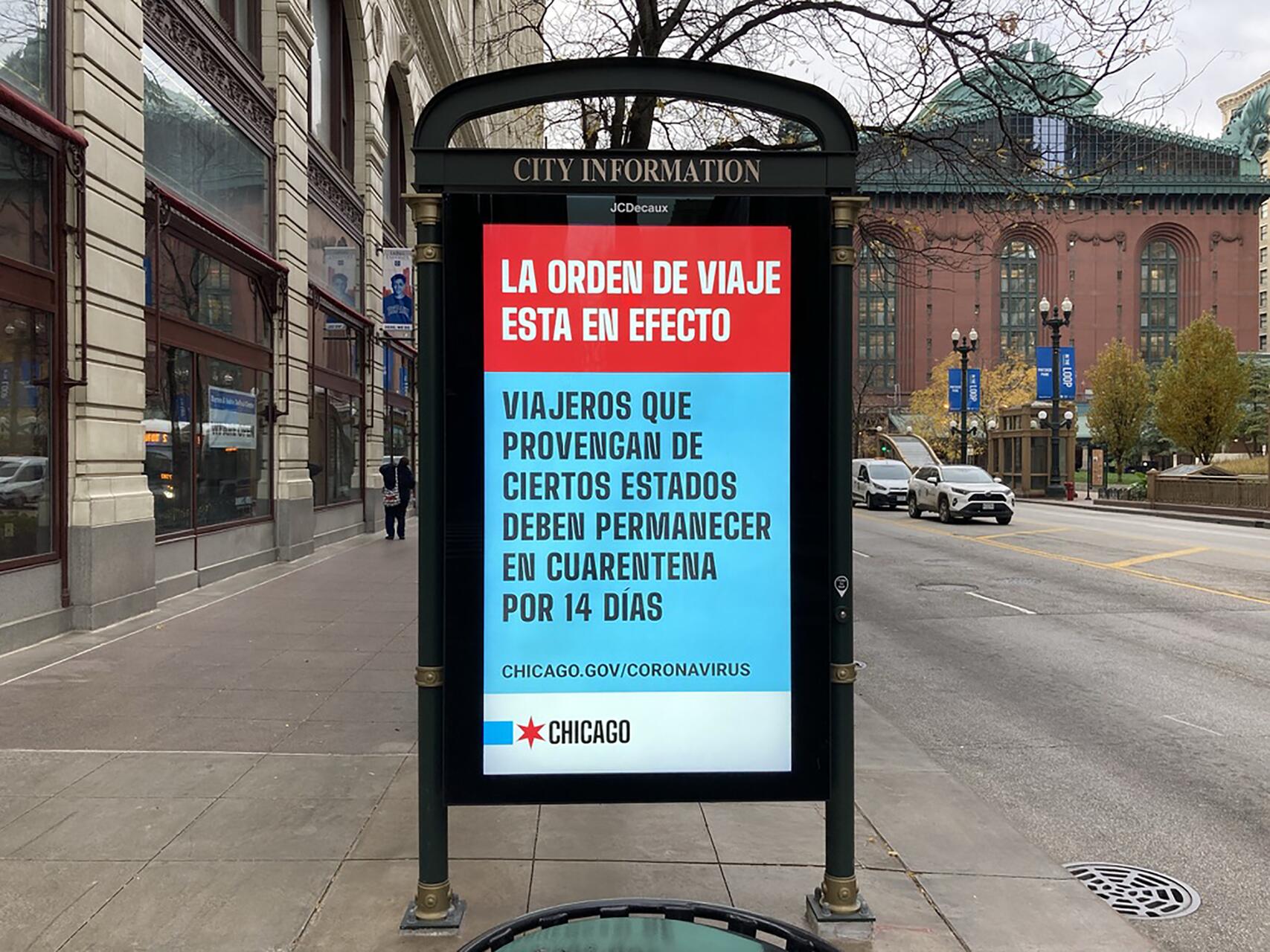Digital city information board on a sidewalk displays a travel order notice in Spanish about quarantine requirements. Street scene shows buildings, trees, and passing cars.