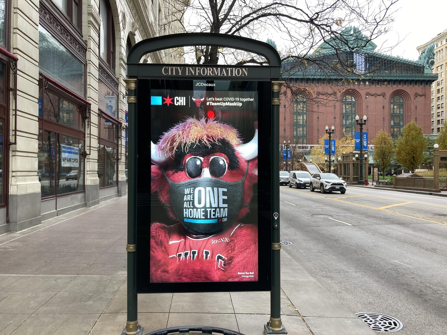 A street advertisement features a mascot wearing a mask with the phrase "We are all one home team," set against a city backdrop with historic buildings.