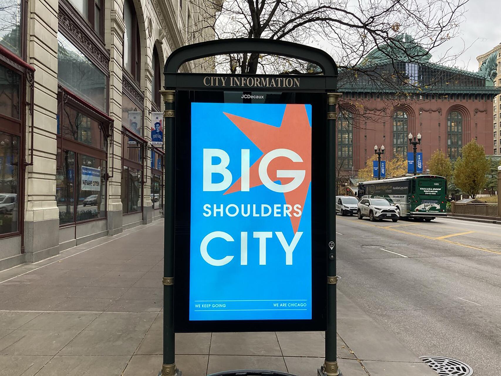 Bus stop advertisement with the text "Big Shoulders City" on a blue background, featuring a red star. Located on a city sidewalk, with street, cars, and buildings visible in the background.