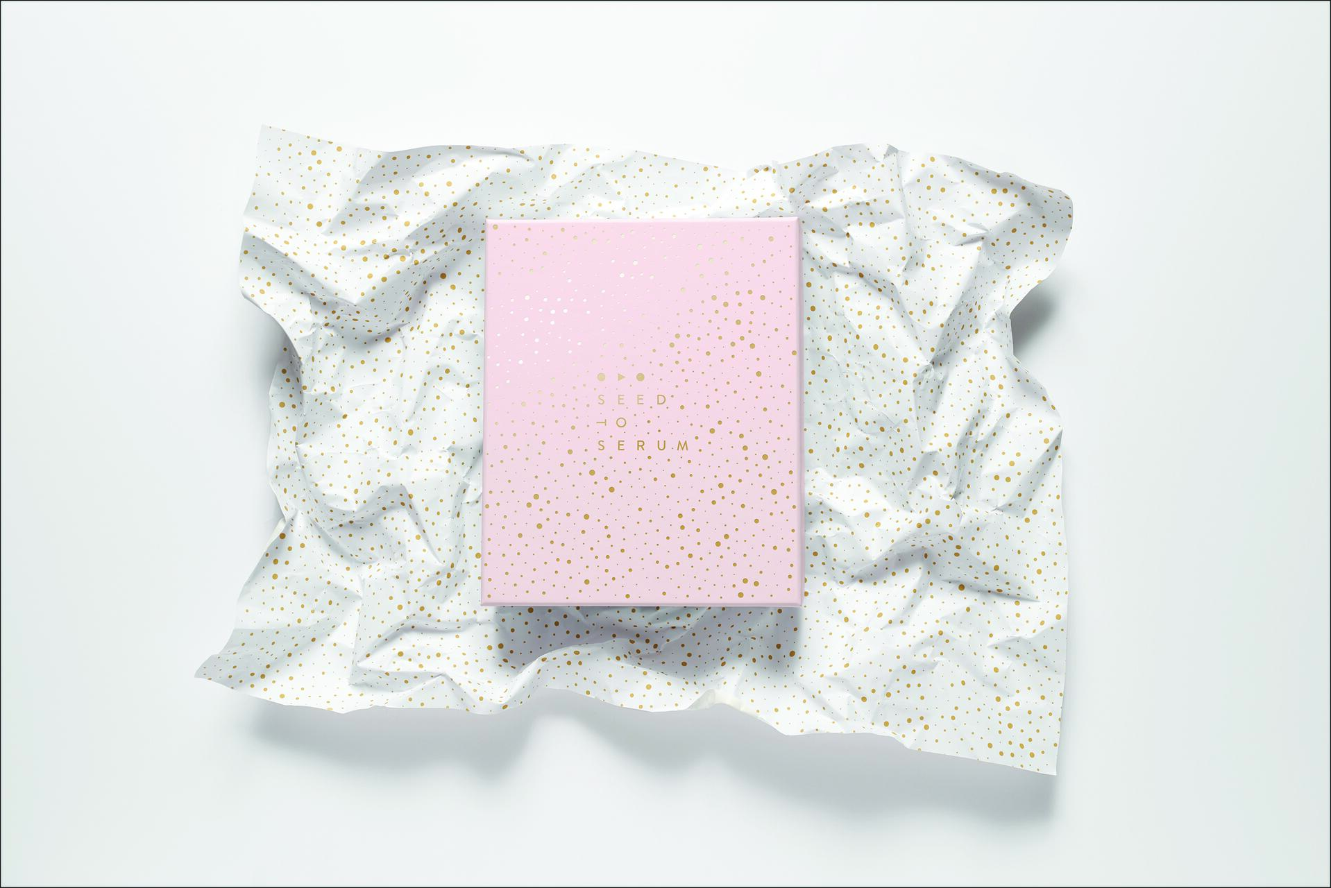 Pink box with gold dots, labeled "Seed to Serum," placed on crumpled white paper with matching gold dots.