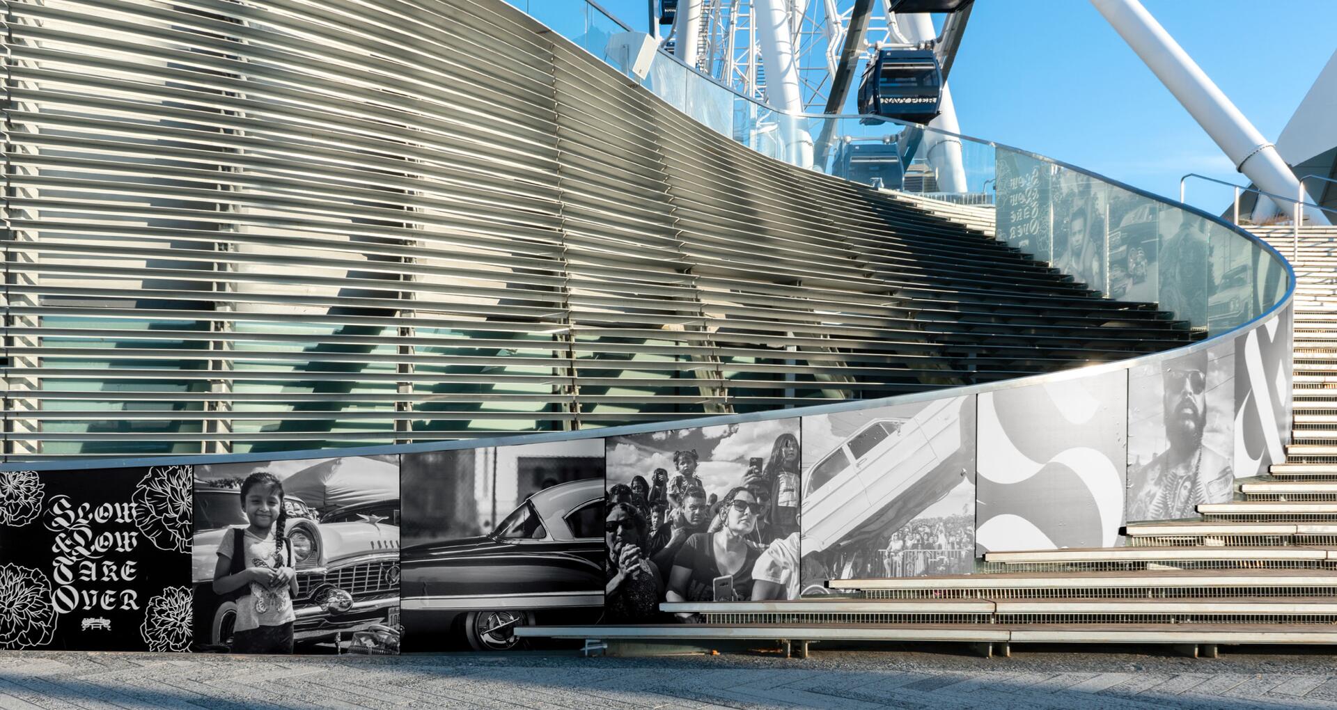 Curved staircase with metal railings, adorned with black and white murals. Background shows the structure of a Ferris wheel under a clear blue sky.