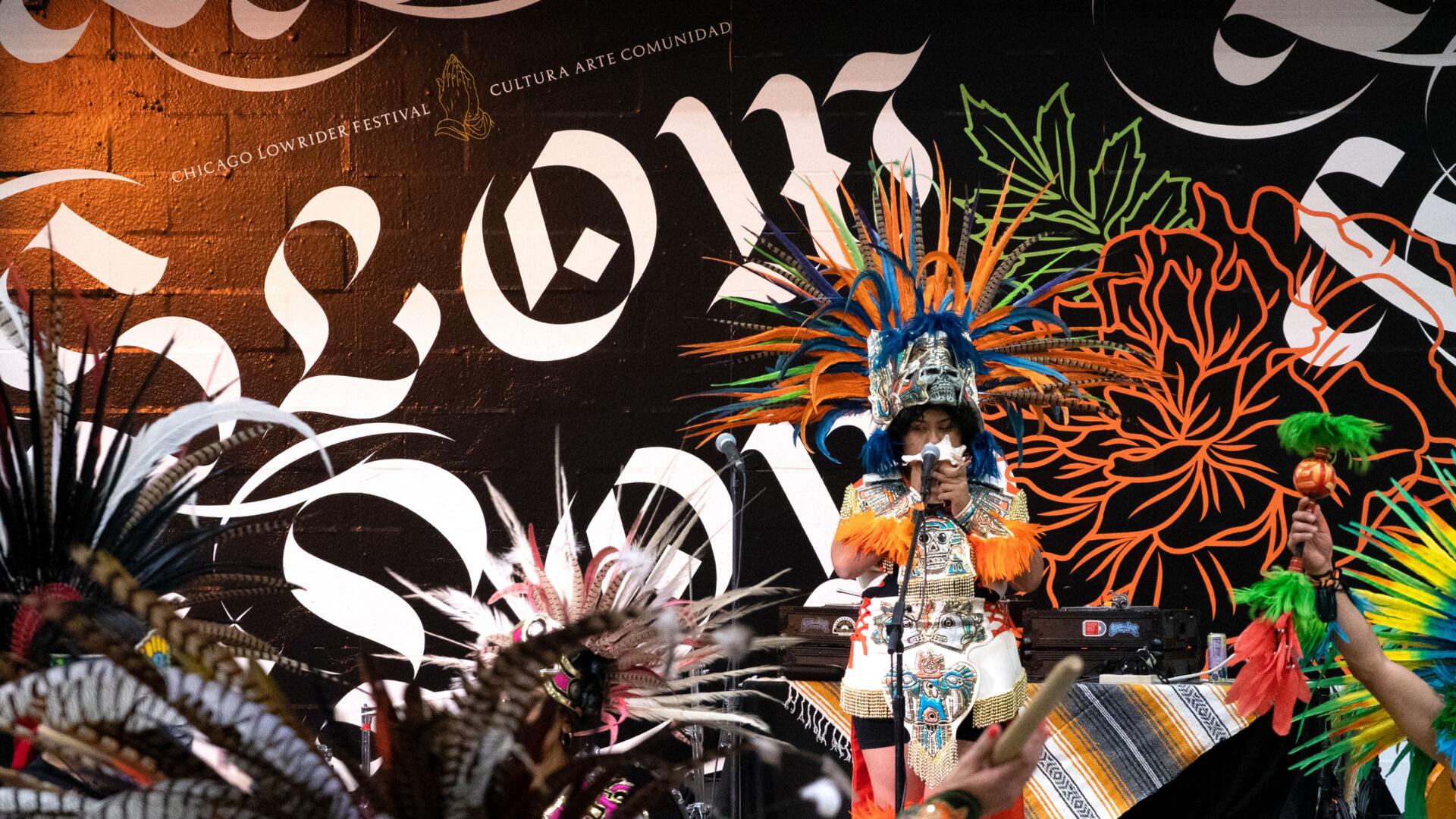 A person wearing a colorful traditional headdress and costume stands on stage holding a microphone, with vibrant murals and "Chicago Lowrider Festival" text in the background. Other performers' feathered headdresses are partially visible.