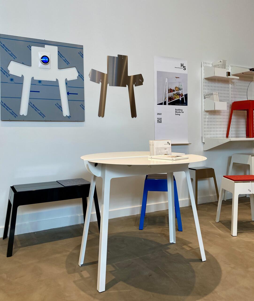 Showroom displays modern furniture prototypes, including a white table, black bench, blue and red stools. Metal cut-outs hang on the wall as design templates. The setting is minimalist with neutral tones.