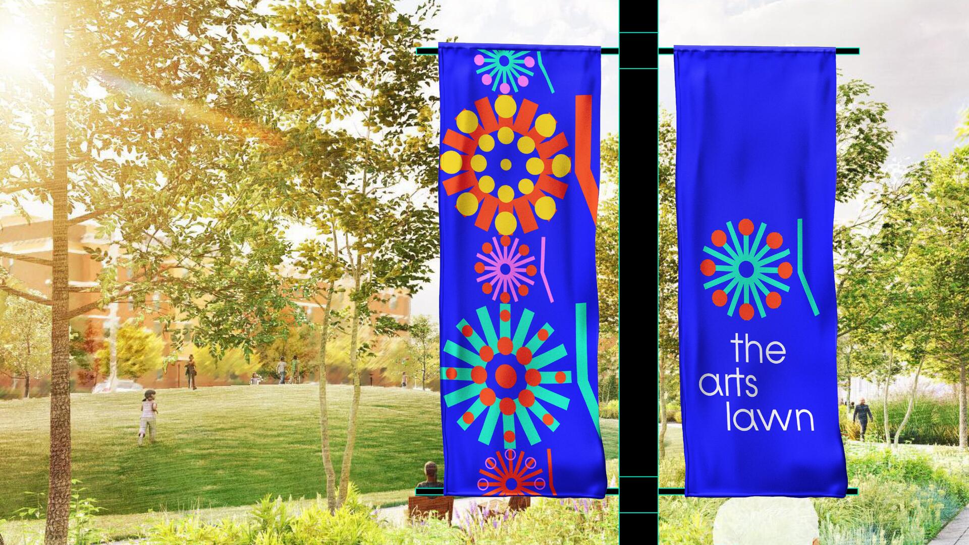 Blue banners with colorful circular designs hang on a pole in a sunlit park setting. People are scattered in the background, and sunlight filters through the trees.