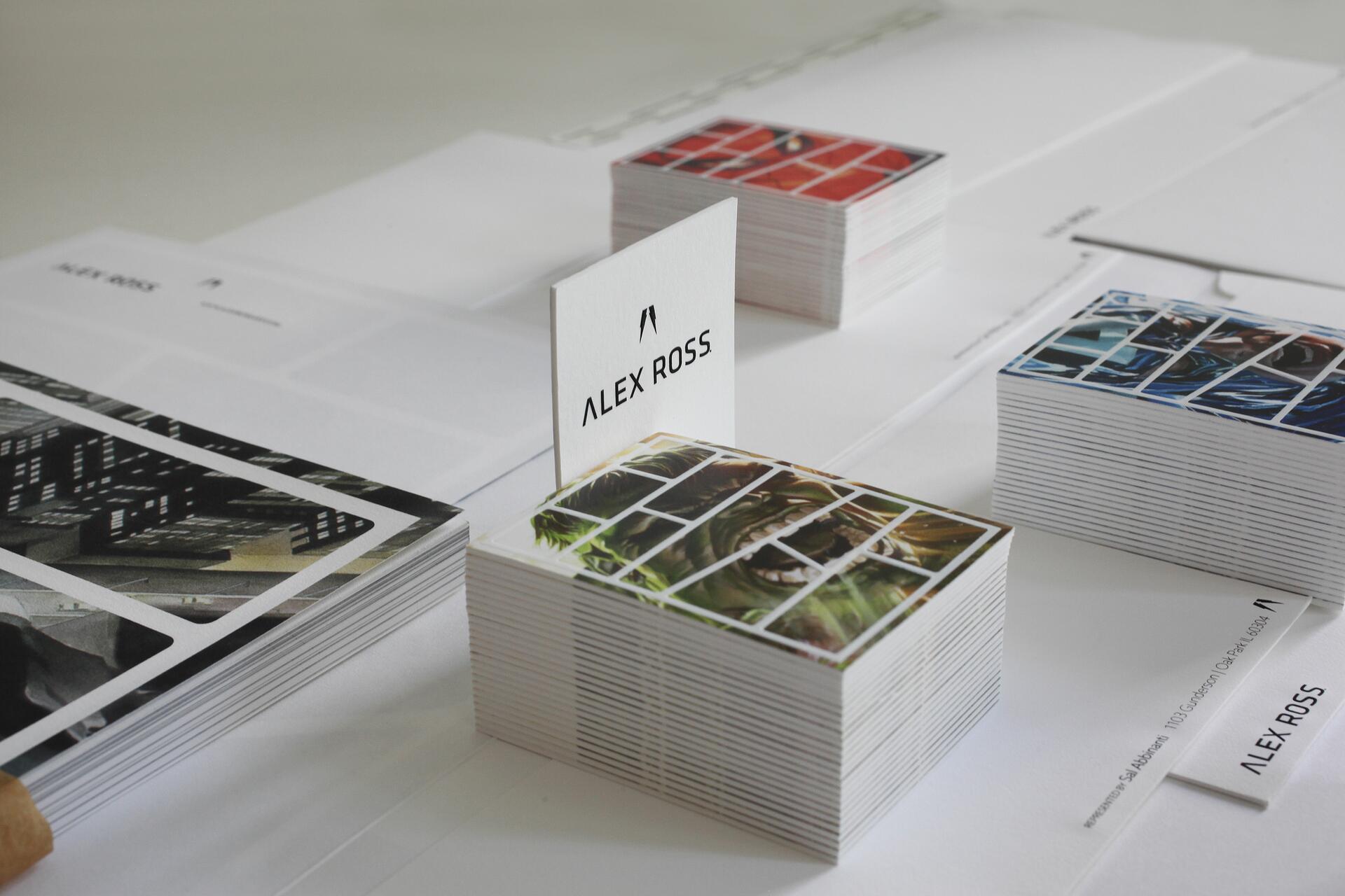 Stacks of art cards arranged neatly on a table, featuring colorful illustrations in orange, green, and blue hues. A visible card reads "ALEX ROSS" in black text, with more stacks in the background.