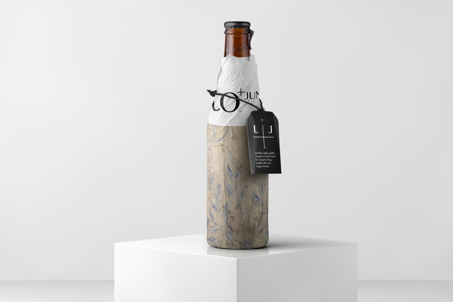 A bottle wrapped in paper with a leaf pattern stands on a white pedestal. A white and brown cap is visible at the top. A black tag with white text is attached to the neck. The background is plain and light gray.