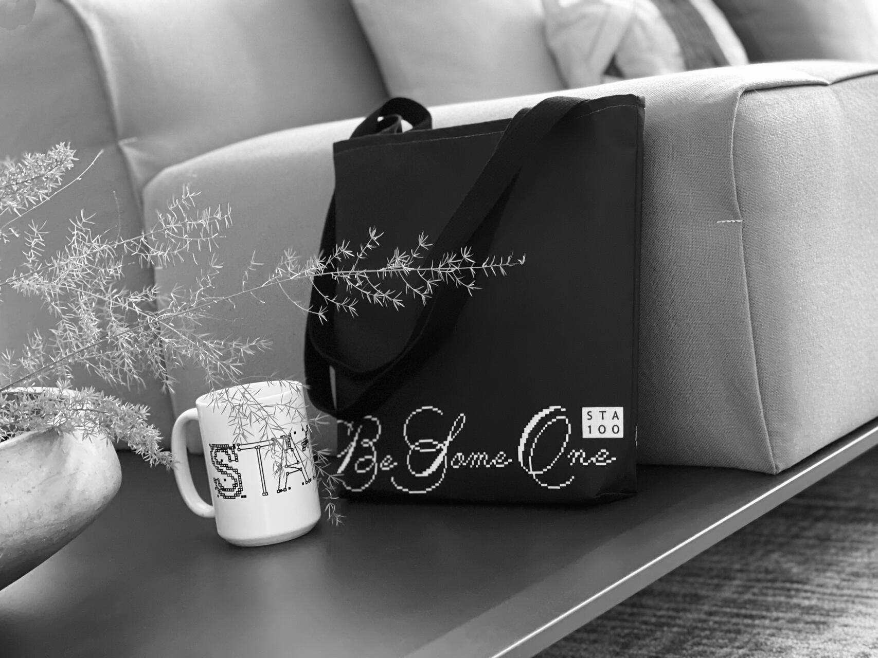 Black and white photograph of a black tote bag with cursive writing, placed on a sofa next to a white mug with bold lettering. A delicate plant in a pot sits beside them on the table.