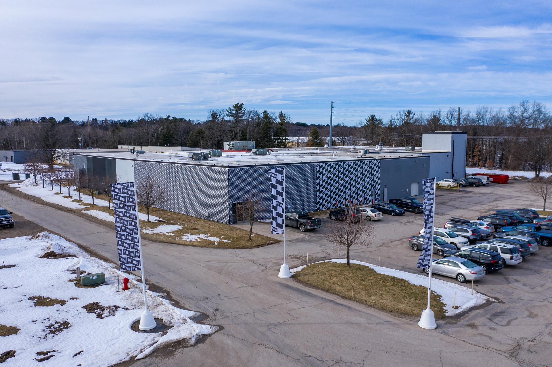 Large industrial building with a geometric facade, surrounded by a snow-dusted parking lot filled with cars. Two tall flags and leafless trees line the driveway. A clear blue sky and distant trees form the backdrop.