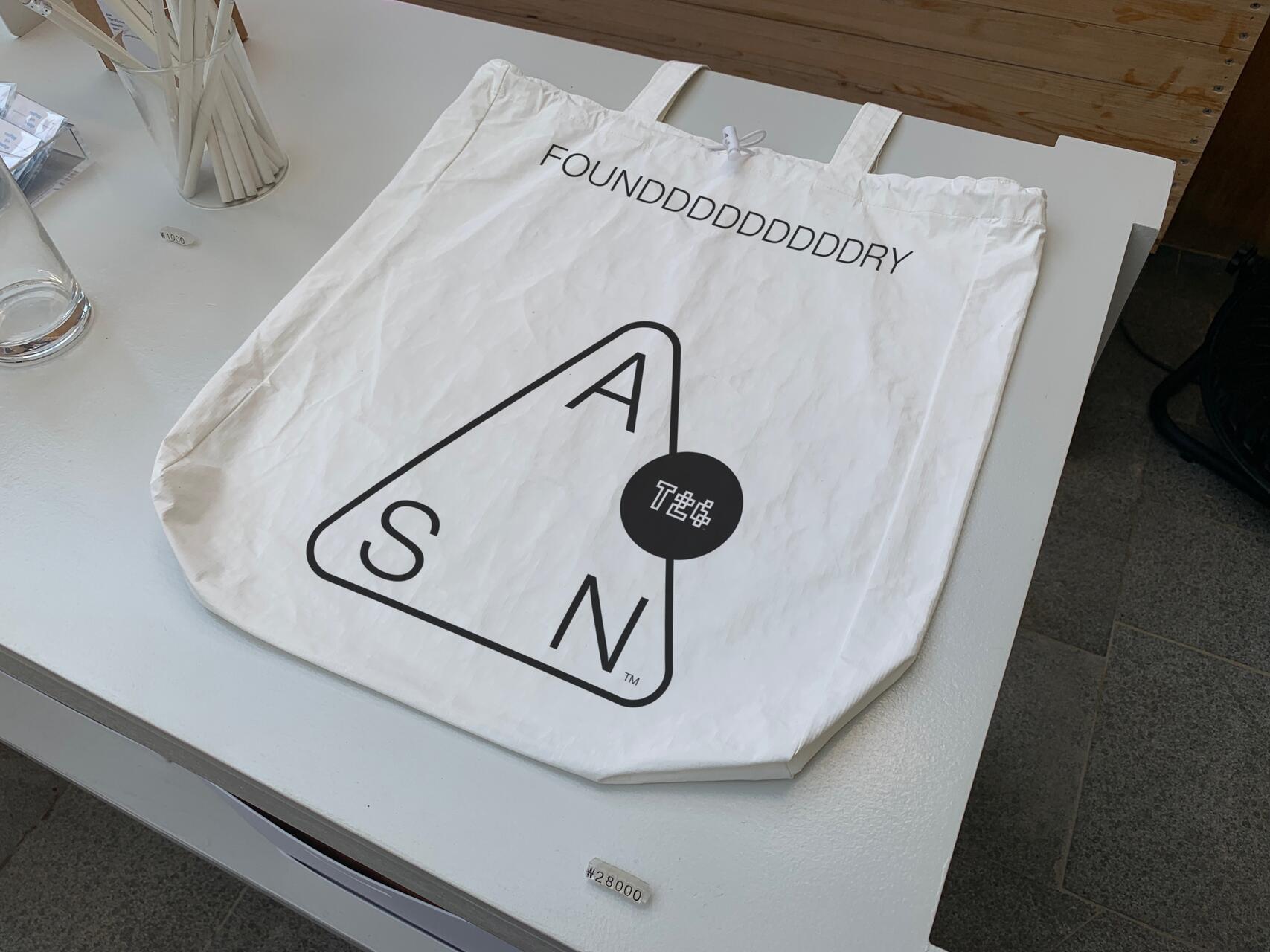 White tote bag on a table with a triangular logo containing the letters A, S, and N, and text reading "FOUNDDDDDDDDDDDDDRY." A glass and other items are in the background.
