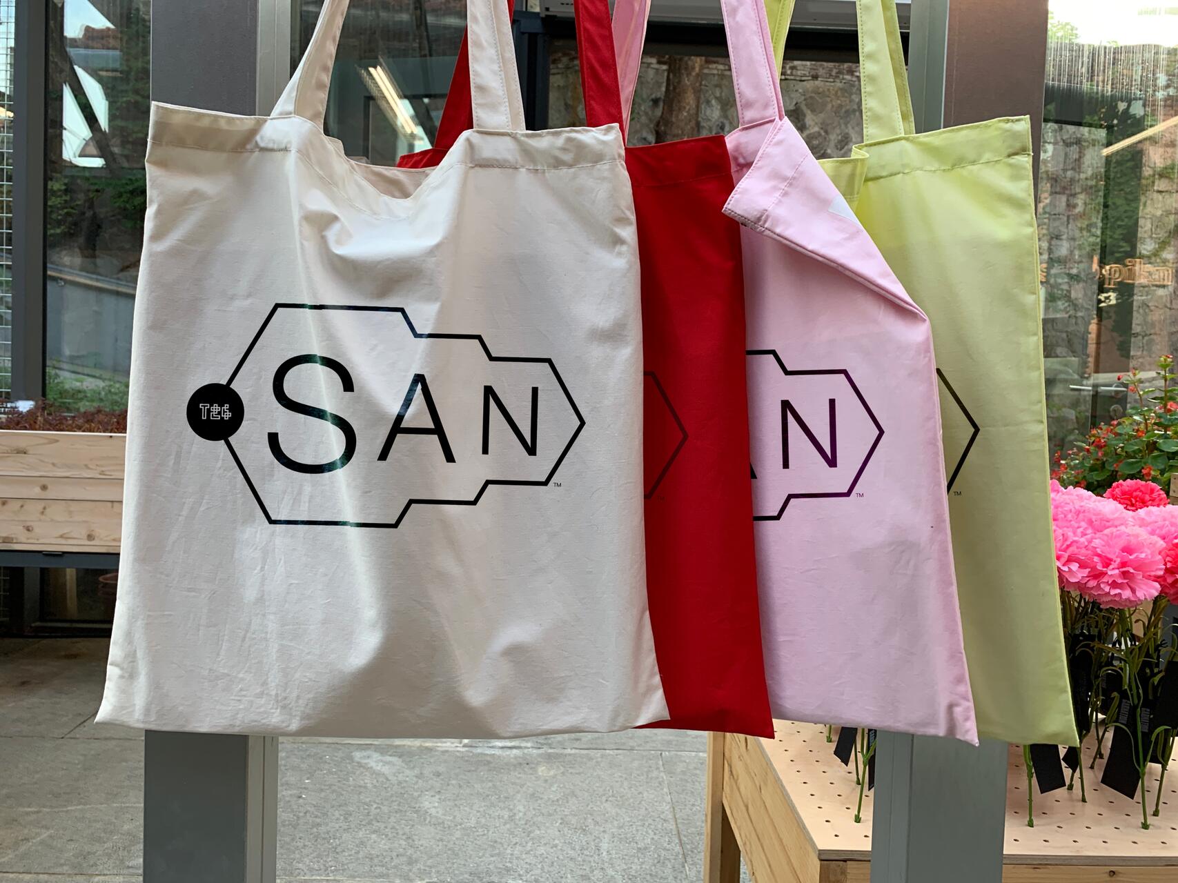Four tote bags hanging in a row, colored white, red, pink, and light green, with the largest white bag prominently displaying a stylized "SAN" logo. The setting appears to be a light-filled shop with flowers in the background.