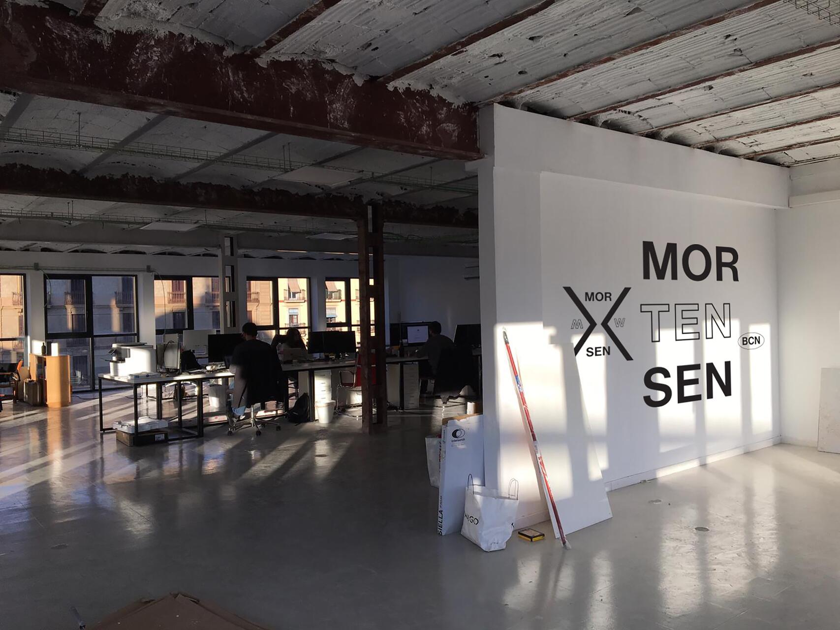 Spacious industrial office with exposed beams, large windows, and a polished concrete floor. Two people work at desks with computers in a sunlit area. Walls feature the text "MORTENSEN."