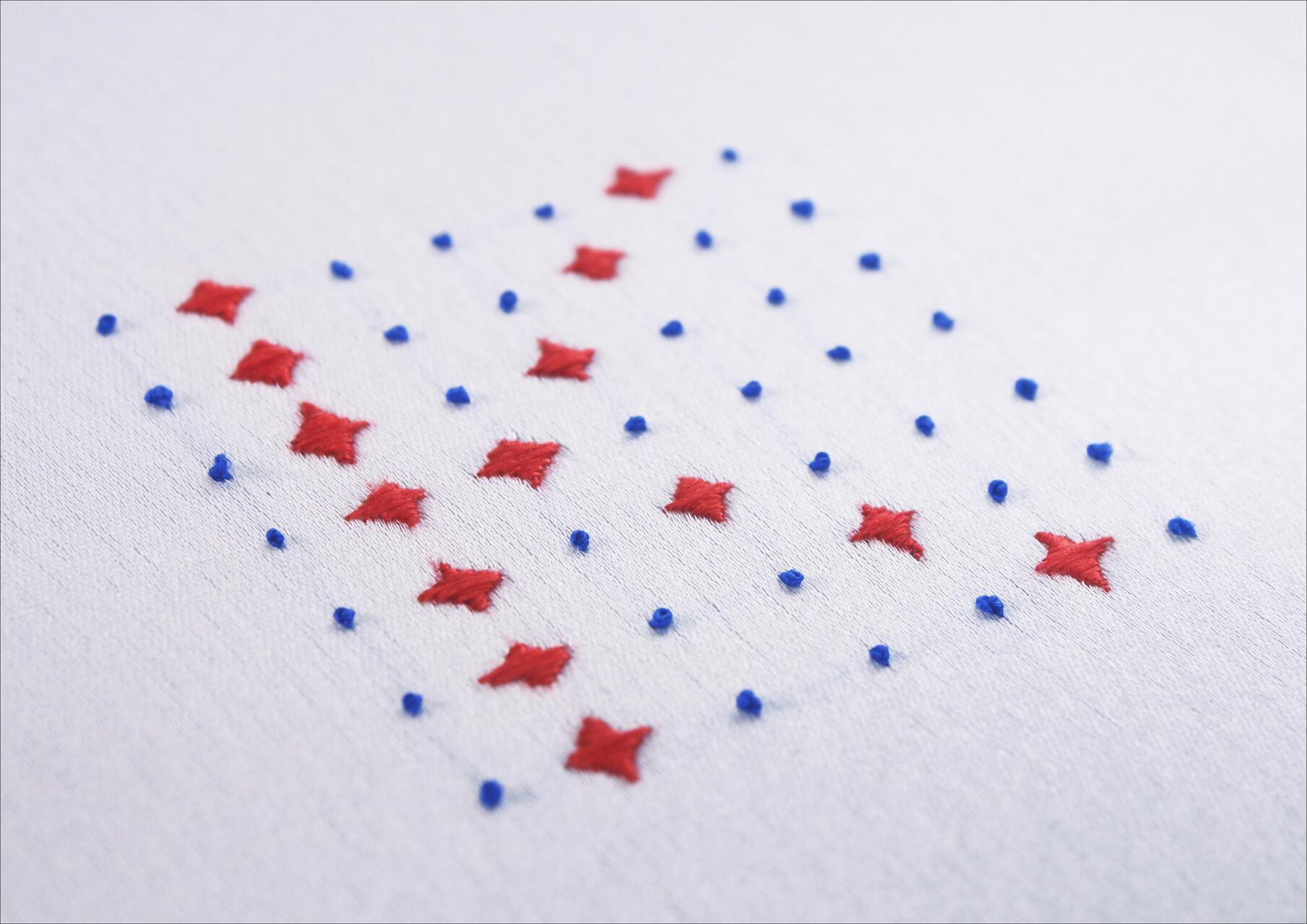 Red diamond shapes and blue dots embroidered on white fabric in a geometric pattern.