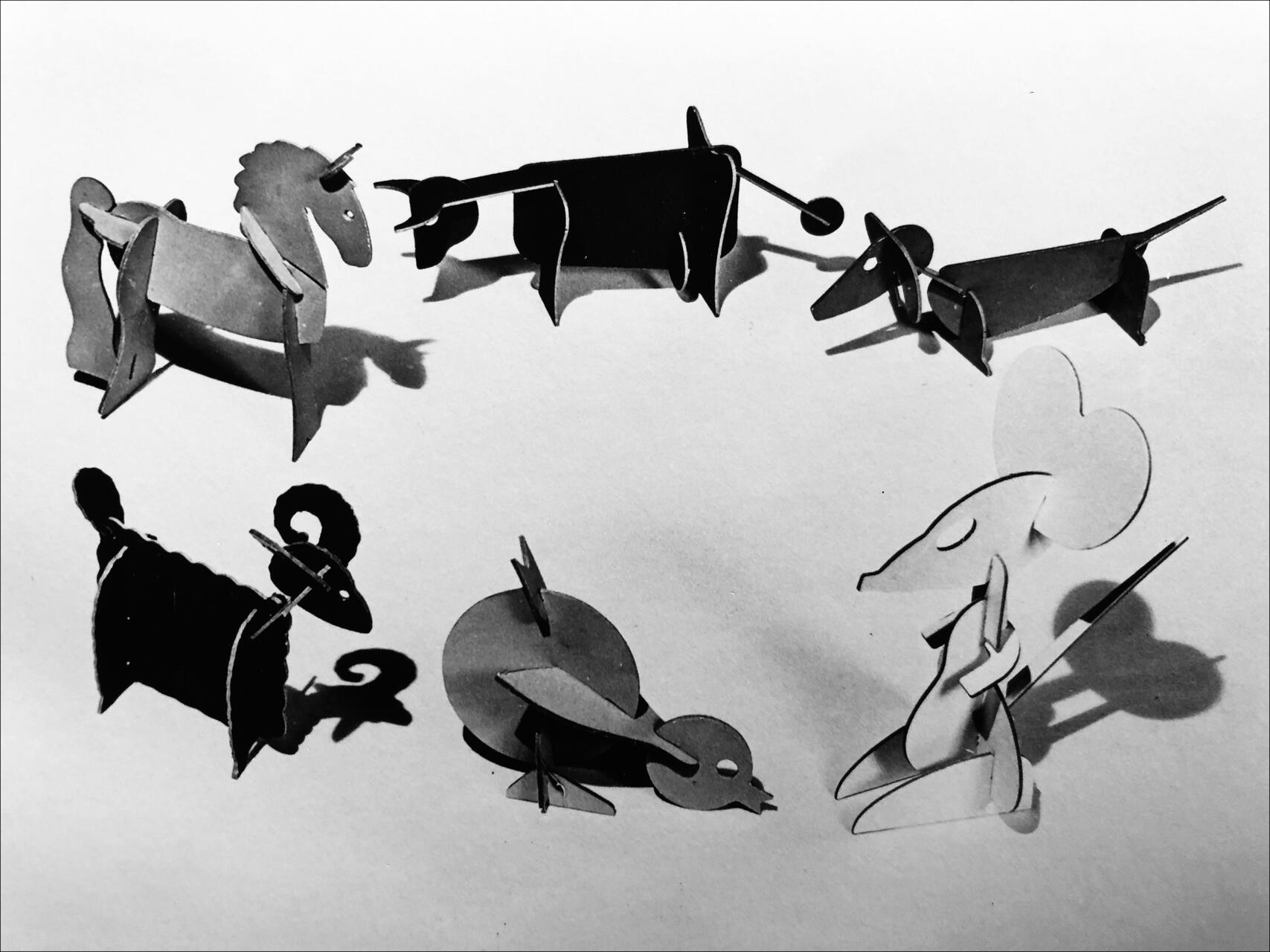 Paper animal models in various shapes including a unicorn, cow, dog, ram, duck, and mouse arranged in a circle on a flat surface, with shadows casting a playful pattern. Black and white photograph.