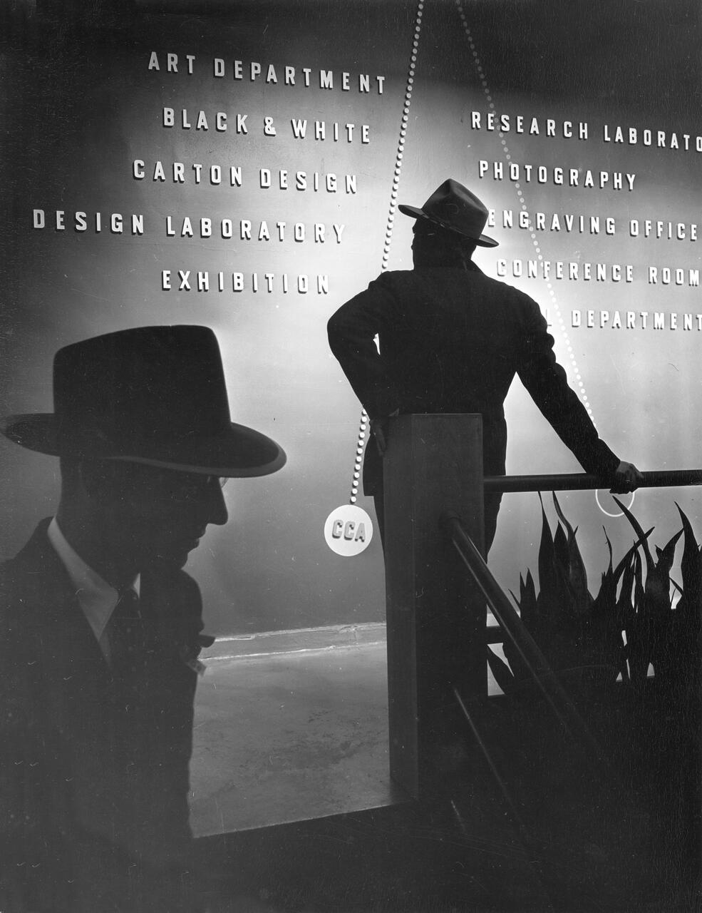 Silhouettes of two men in suits and hats stand in front of a wall with labeled departments, with one man's reflection visible. The ambient lighting casts dramatic shadows, creating a mysterious atmosphere.