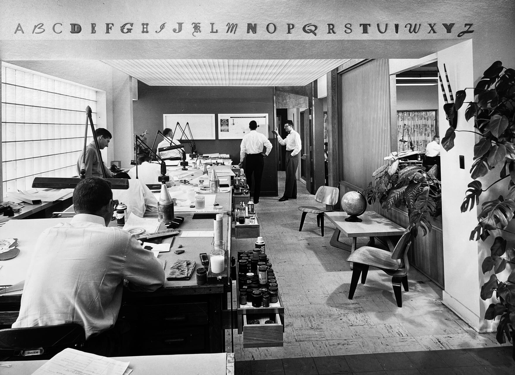 Black and white photo of a mid-20th-century office with a curved desk covered in papers and art supplies. Two men are working while two others converse by a wall chart. Decorative plants and a globe are in the foreground, with alphabet letters above.