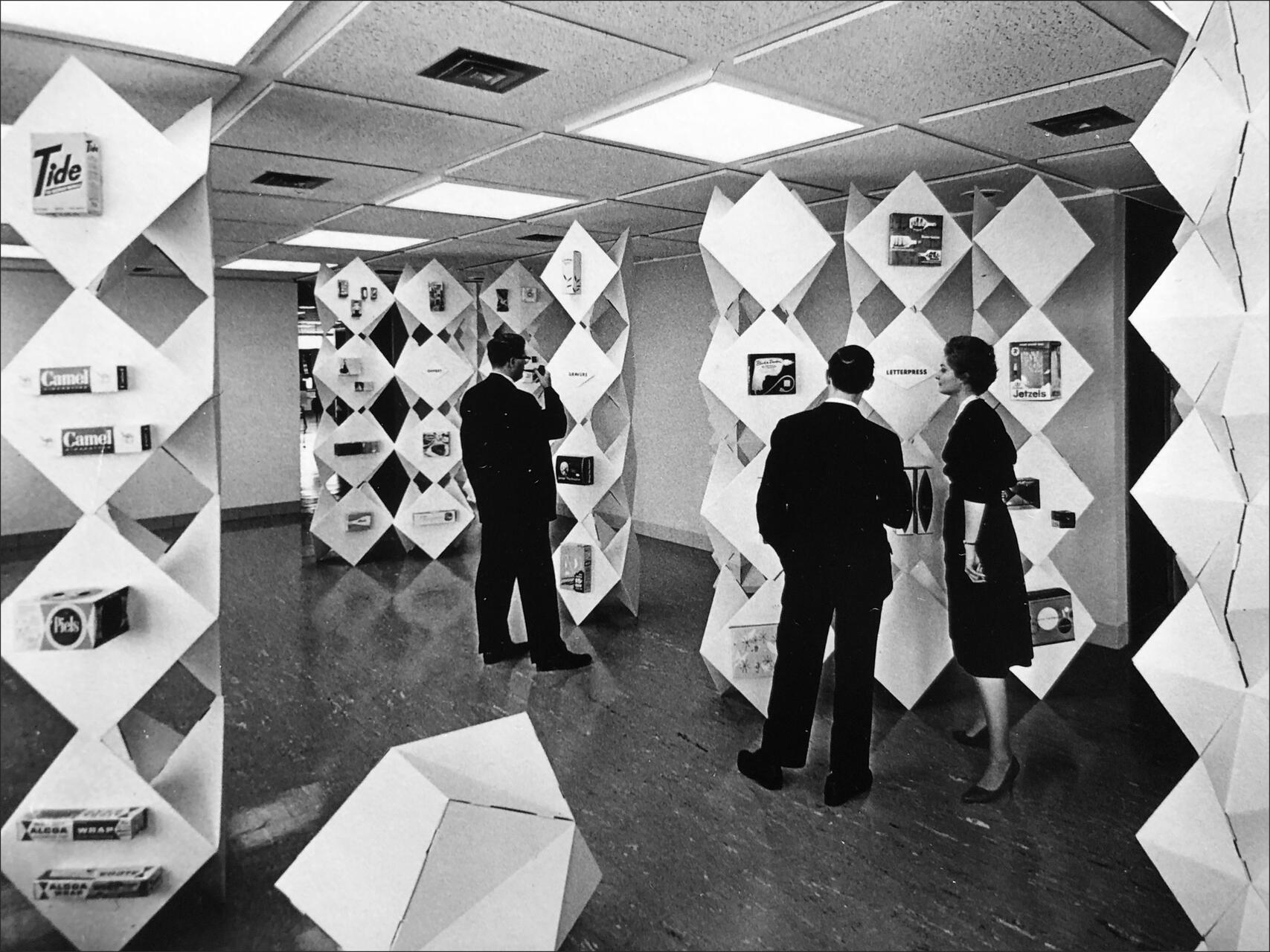 People in a gallery-style room with geometric white lattice shelves displaying various product boxes, including Tide and Camel, under grid-like ceiling lights.