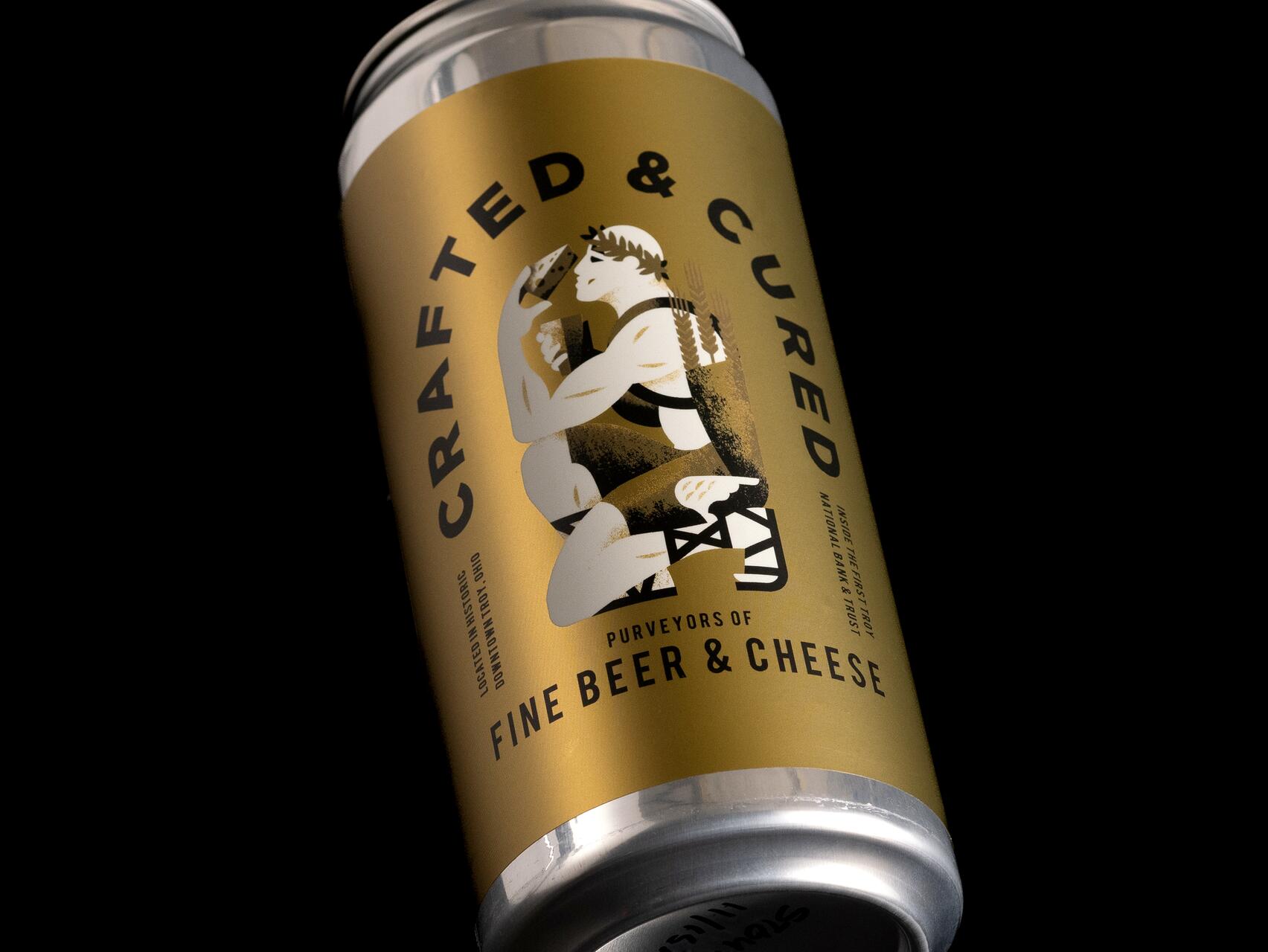 Golden can with the label "Crafted & Cured" featuring an illustrated figure holding a mug, surrounded by the words "Purveyors of Fine Beer & Cheese." The design includes black text and graphics on a metallic background, set against a black background.