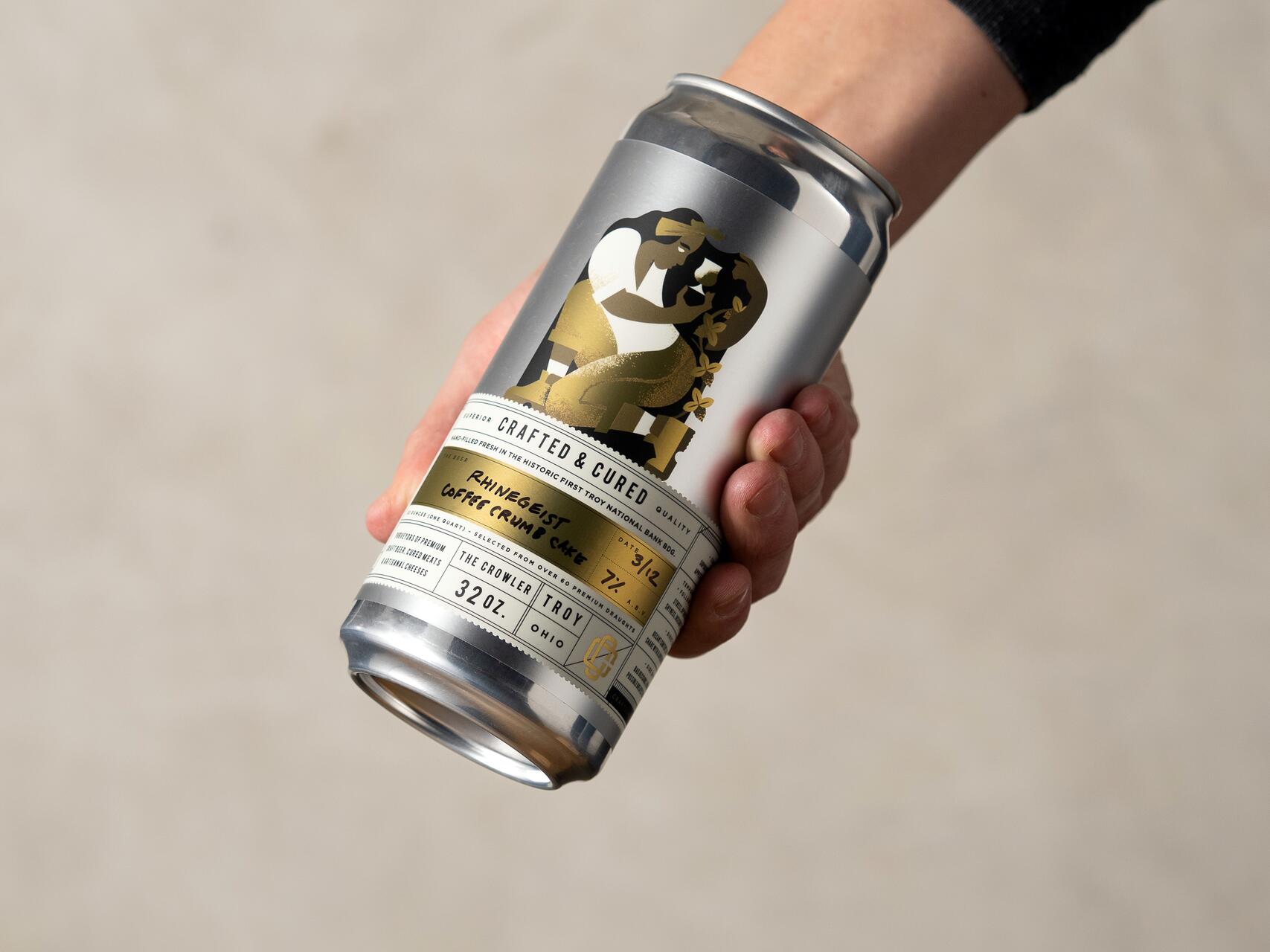 A hand holding a silver can labeled "Rhinegeist Coffee Crumb Cake," featuring a stylized gold and black illustration. The can is tilted, revealing details of the design, with a soft gray background.
