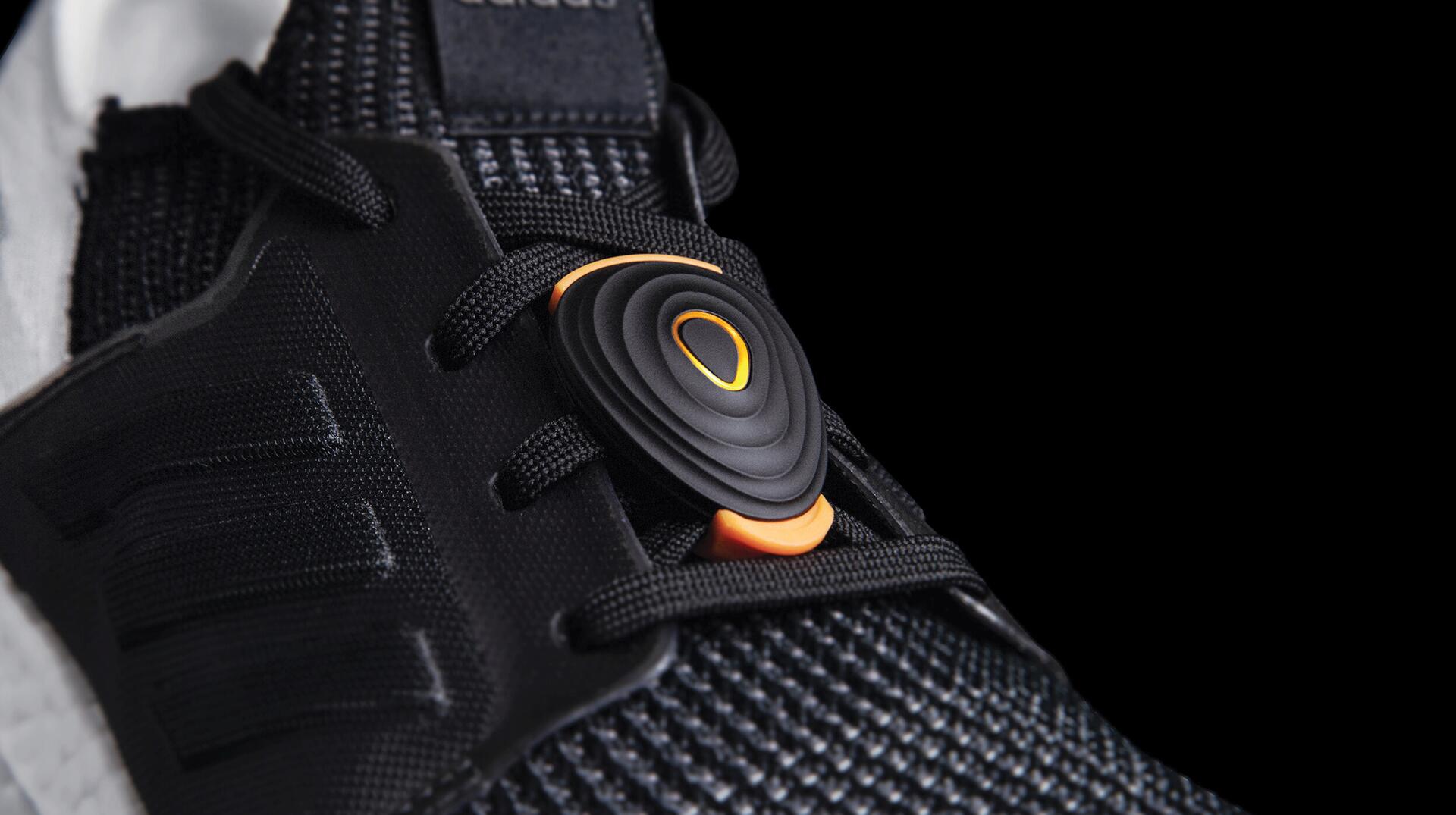 Black sneaker with textured fabric and a circular black and orange clip on the laces, set against a black background.