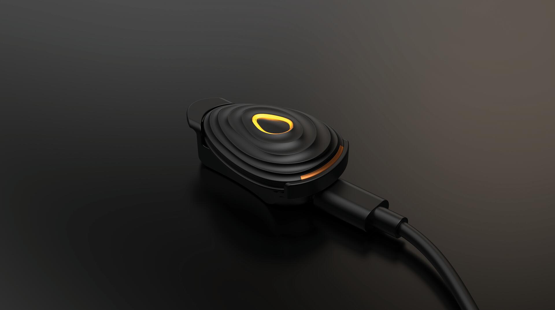 Black electronic device with concentric circular ridges and a glowing orange accent, connected by a cable, on a dark surface.