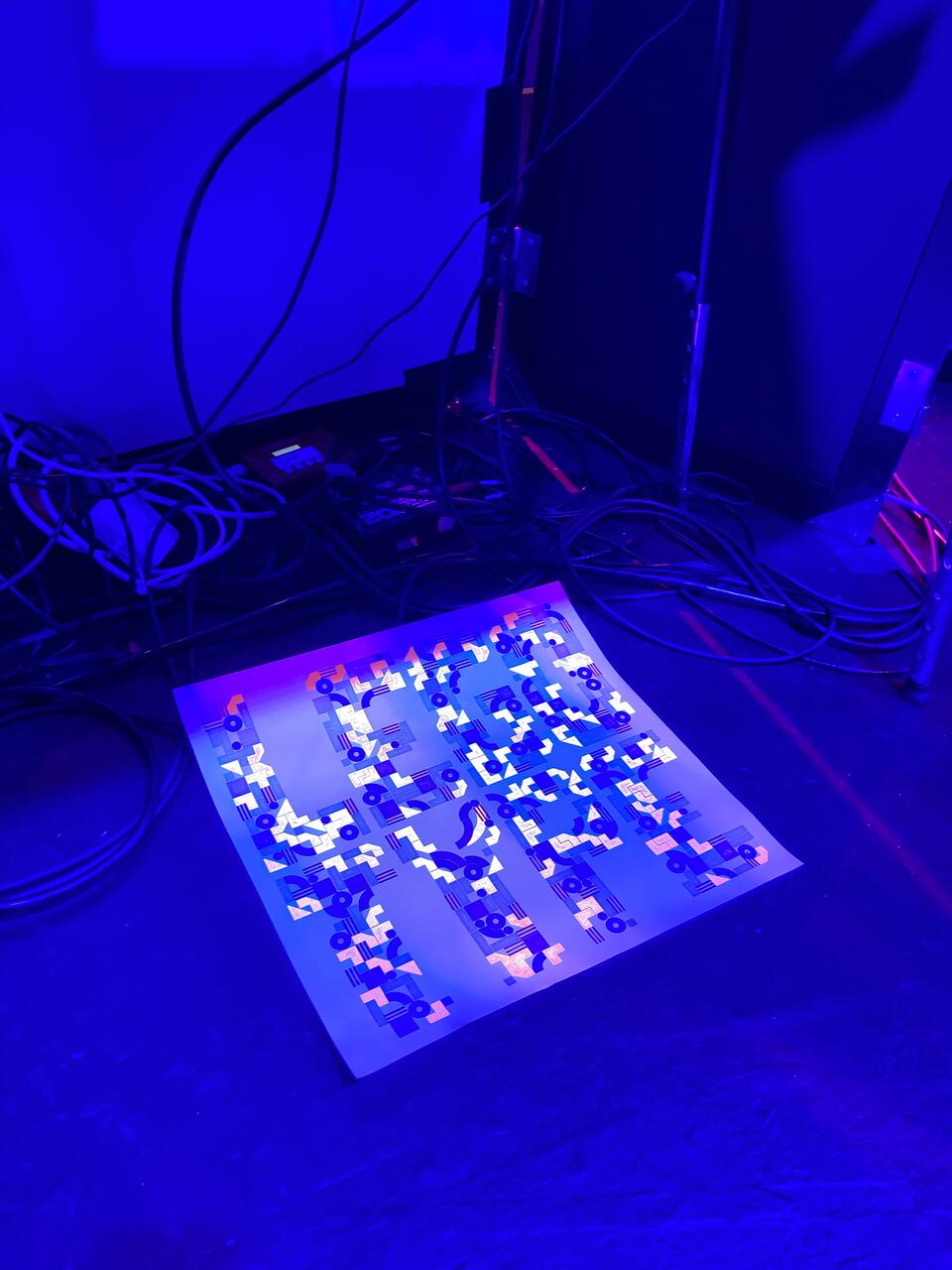 Abstract graphic artwork on the floor, illuminated by blue light, surrounded by cables and electronic equipment.