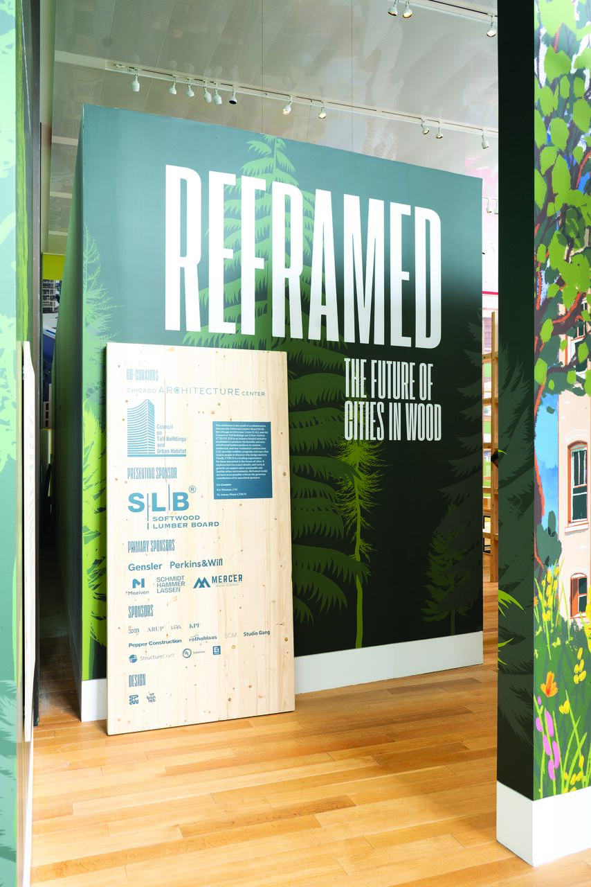 Exhibit display with "Reframed: The Future of Cities in Wood" text on a forest-themed wall. A wooden board lists event sponsors, set in a brightly lit gallery with hardwood floors.