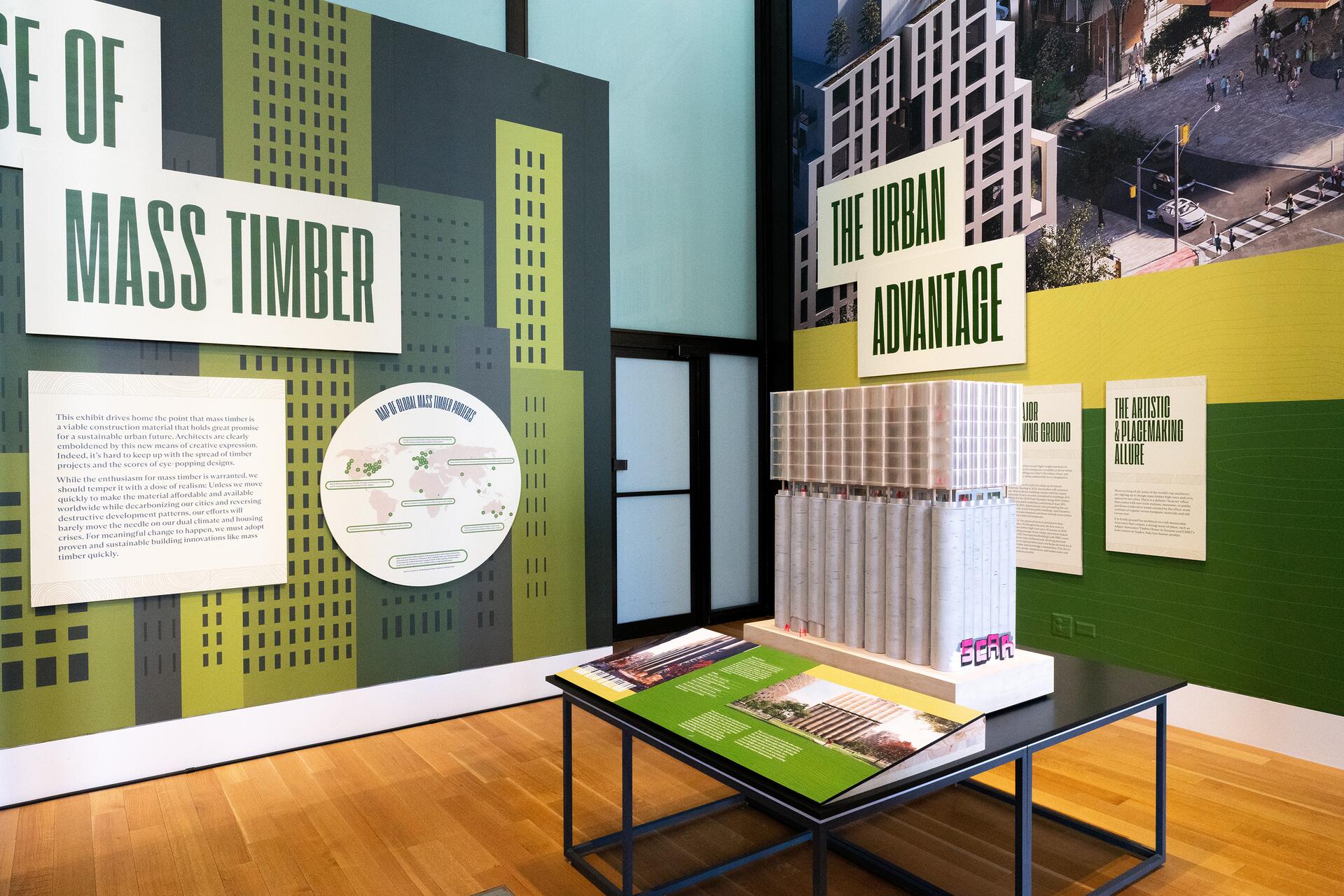Exhibition display featuring informational panels about mass timber construction and a scale model of a tall building. The setting includes graphic representations of cityscapes and text discussing timber's urban benefits.