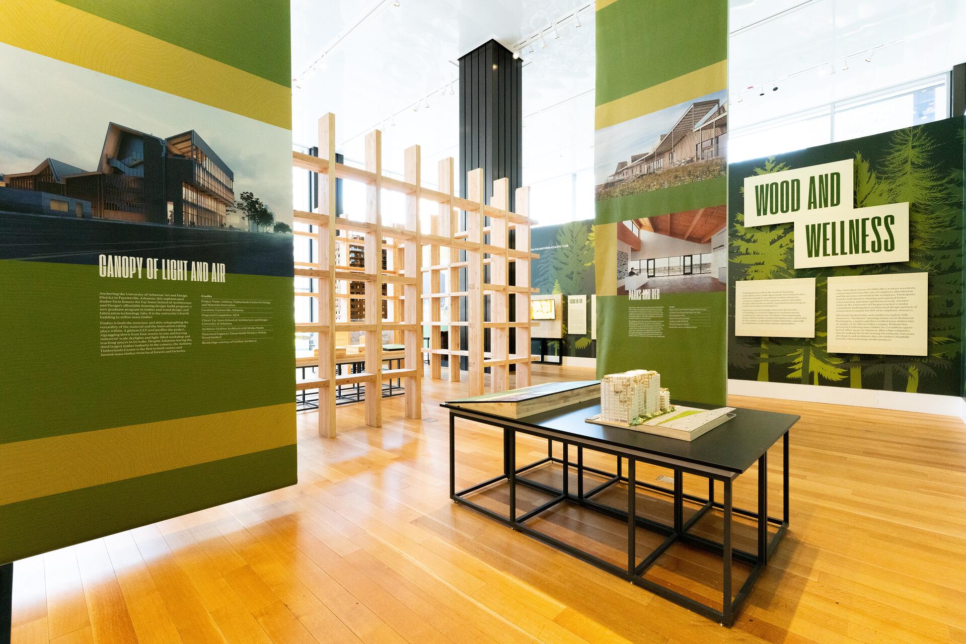 Exhibition room with green panels displaying architectural designs and text, wooden structures, and a table with a scaled building model. Wood flooring and bright lighting create a modern ambiance.