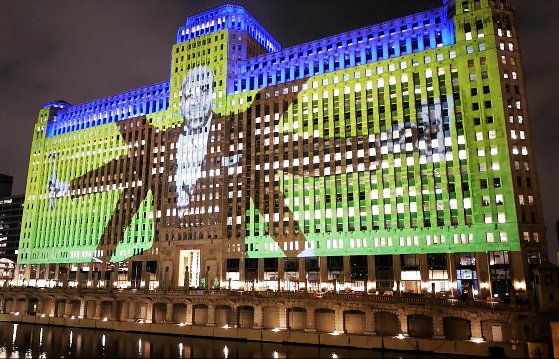A colorful light projection on an elaborate building at night shows a giant figure against a backdrop of blue, green, and yellow. The building's windows create a grid pattern, reflecting in a nearby body of water.