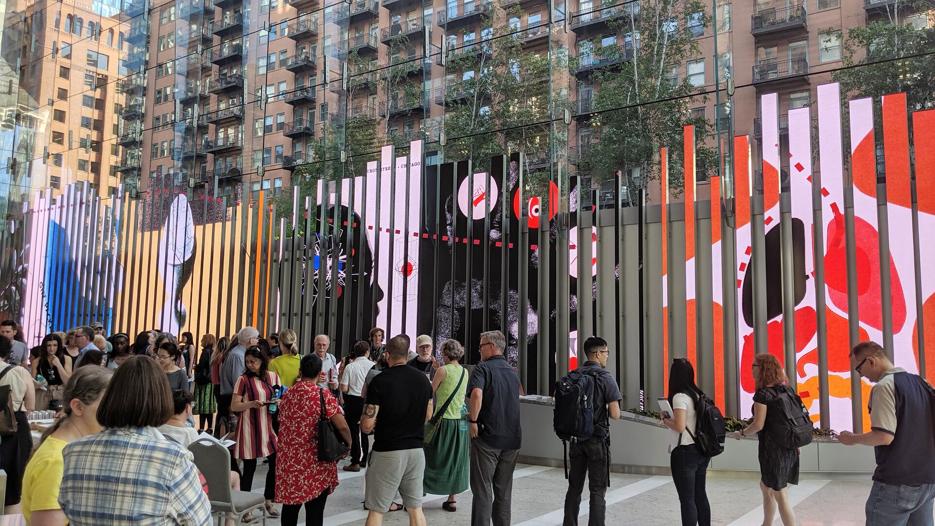 Crowd gathers in front of a large, vibrant digital art display with vertical panels, showcasing colorful abstract images. Urban setting with a backdrop of glass and brick buildings.