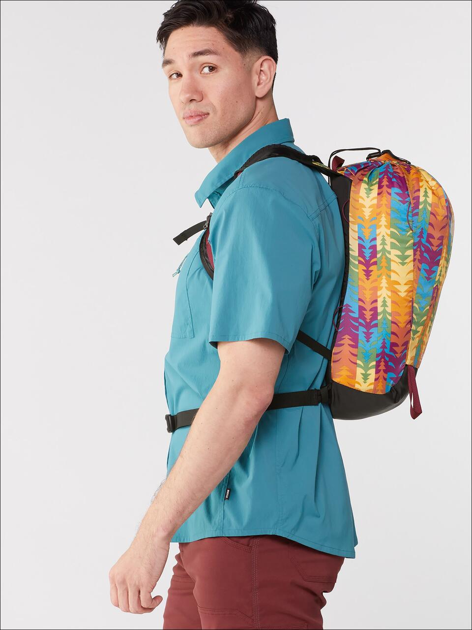Man wearing a teal shirt and maroon pants, carrying a colorful backpack with a bright pattern, looking over his shoulder against a plain background.