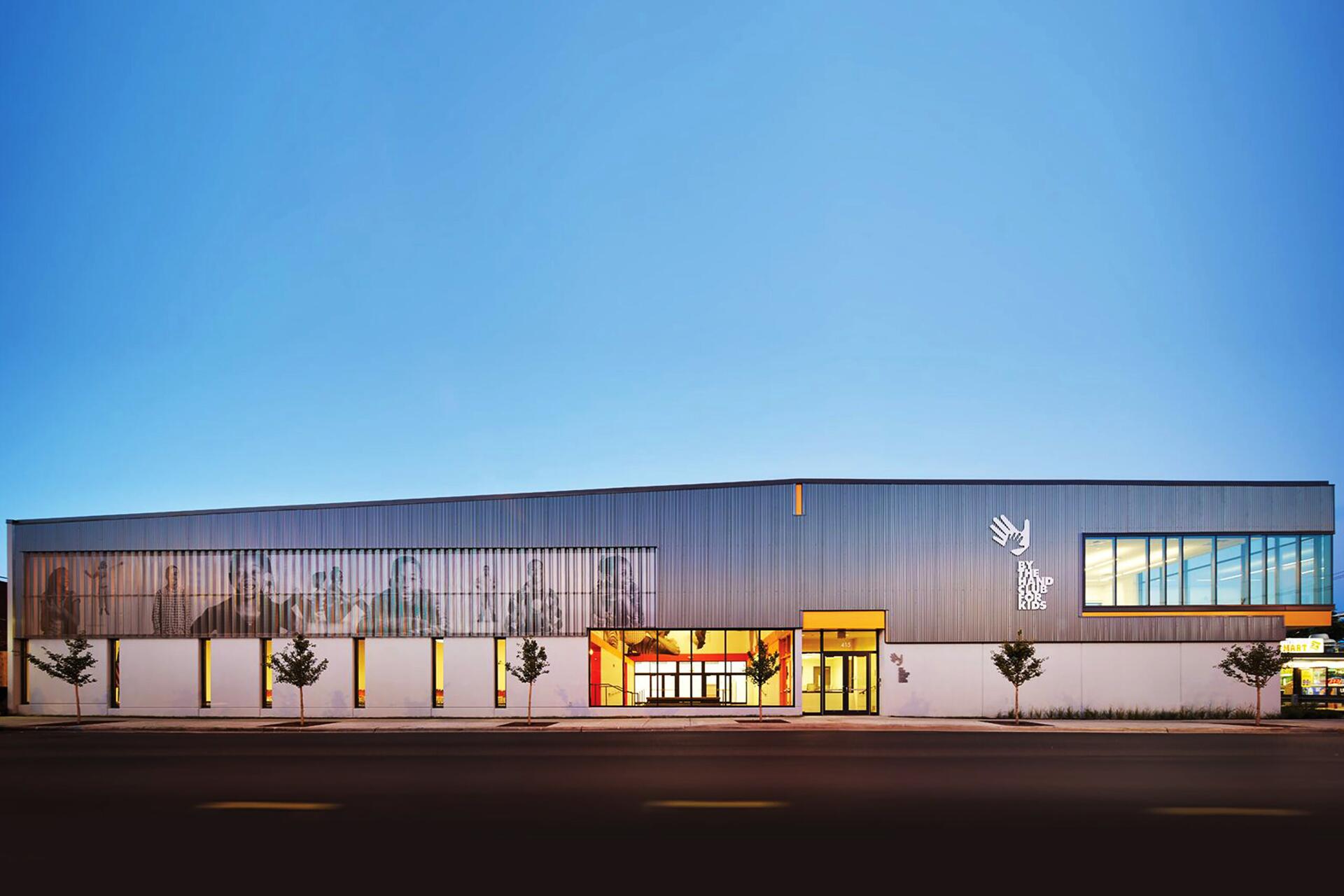 Modern building with a sleek, elongated facade, featuring large windows and a transparent section. The exterior is lit warmly from within, framed by a clear sky. Small trees line the sidewalk.