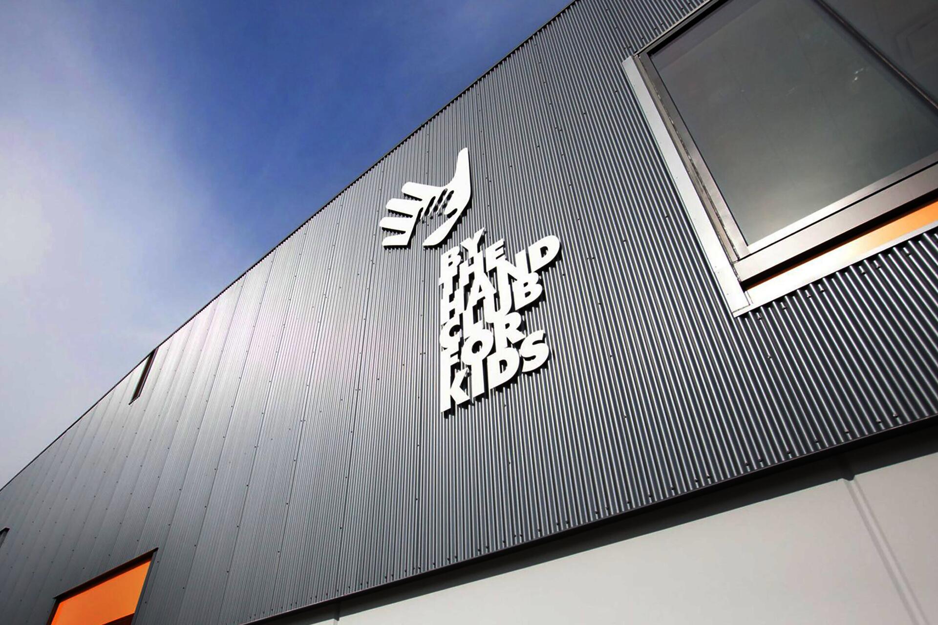 Modern building facade with a corrugated metal texture, featuring a large window and white signage shaped like a hand. The sky is clear and blue.