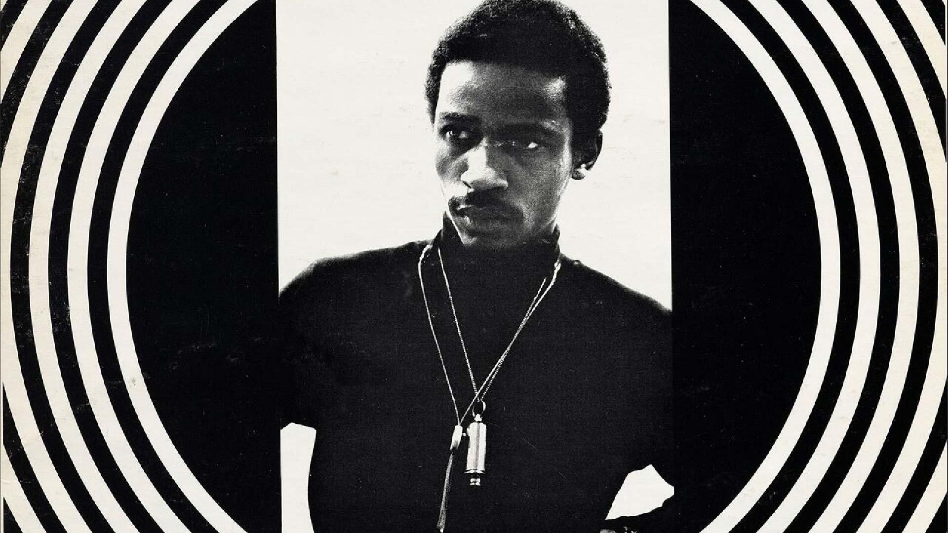 Black and white photograph of a man with a serious expression, wearing a dark turtleneck and necklaces, framed by concentric black and white circles.