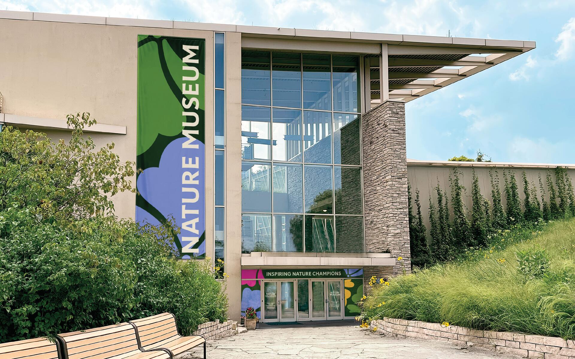 Nature museum entrance with large glass windows and a colorful vertical sign. Lush greenery and a pathway with benches in the foreground. Clear blue sky above.