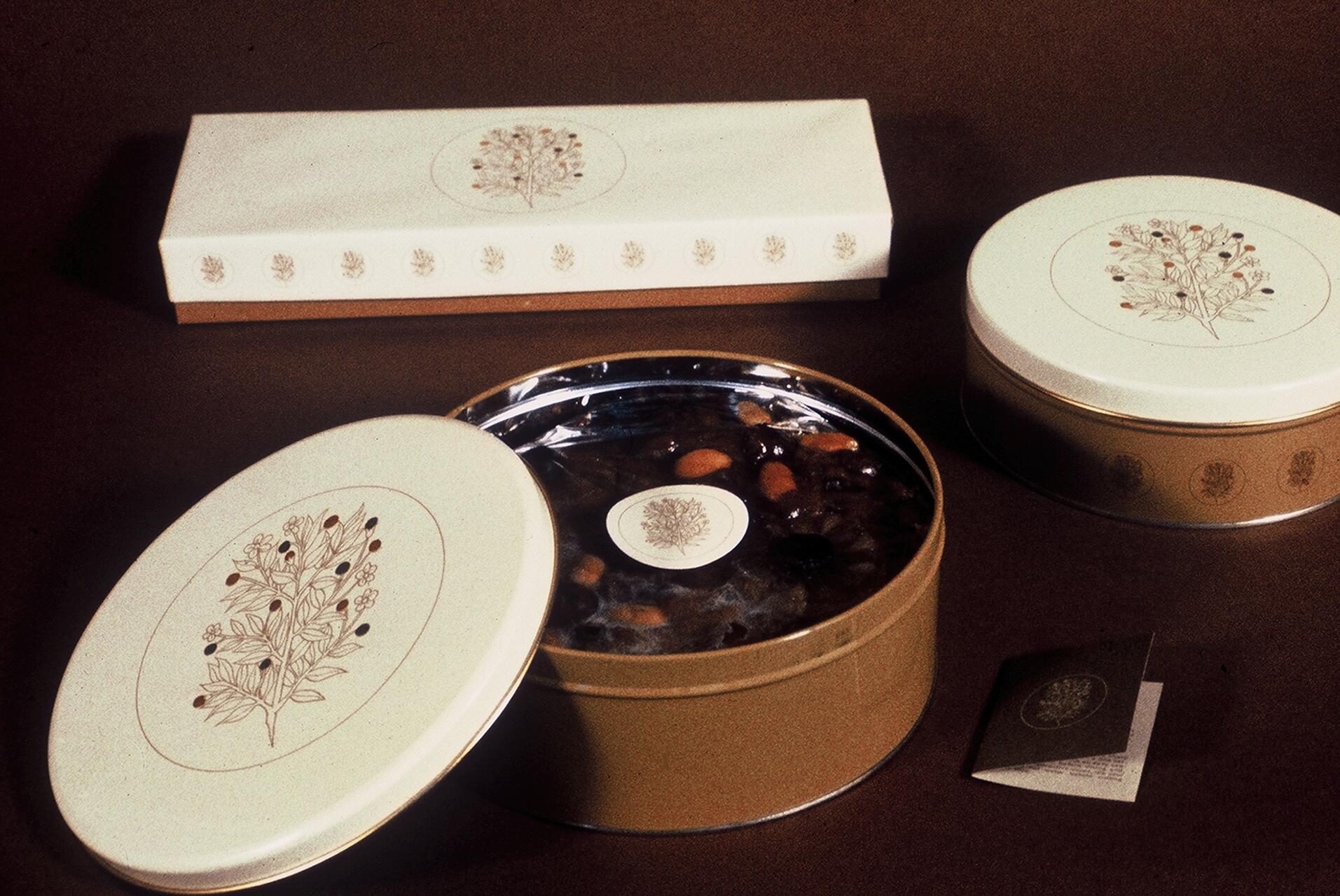 Round and rectangular cream-colored tins with botanical designs, one open to reveal assorted nuts and fruitcake. Brown background.