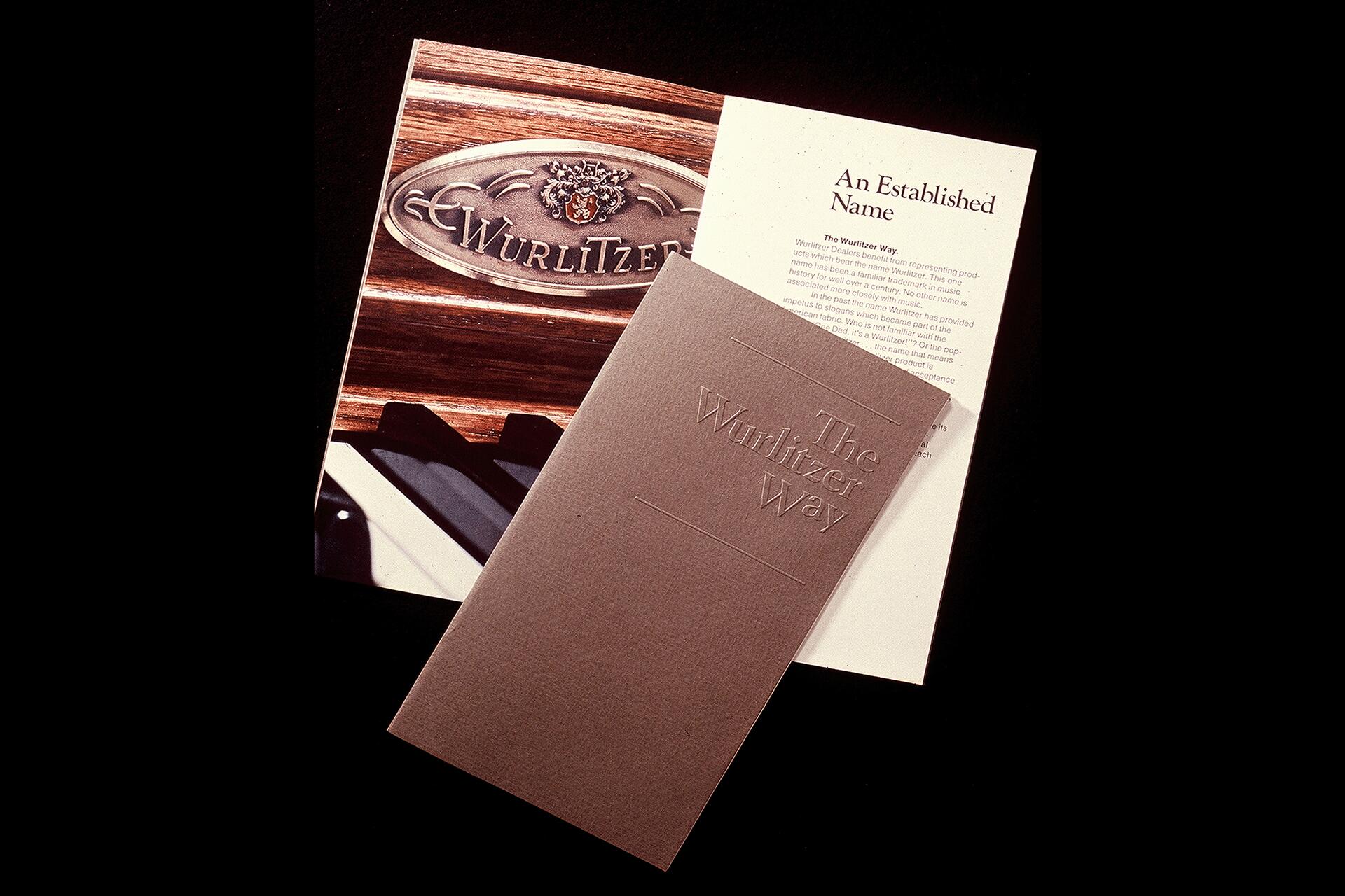 Brochure cover titled "The Wurlitzer Way" with embossed text, laid over a page featuring the Wurlitzer logo against a wood texture and piano keys.