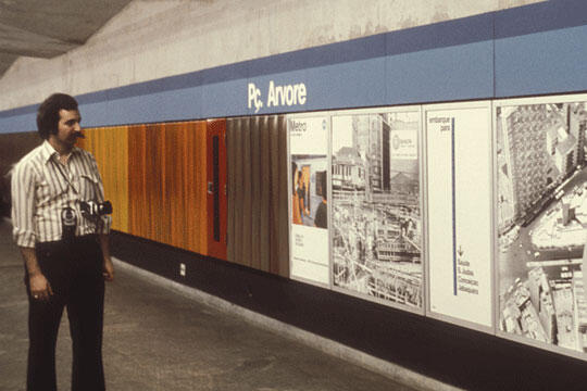 A man with a camera around his neck stands in a subway station, looking at a wall with colorful panels and various architectural posters. The signage above reads "Pc. Anvore."