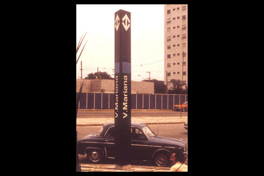 Tall sign in an urban area with "V. Mariana" text, an old black car parked nearby, and a skyscraper in the background under a clear sky.