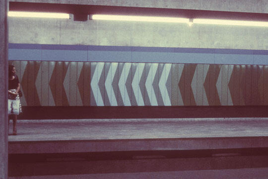 A dimly lit subway platform with a geometric pattern of large, alternating chevrons in shades of brown and white on the wall. A person stands on the left side of the platform.