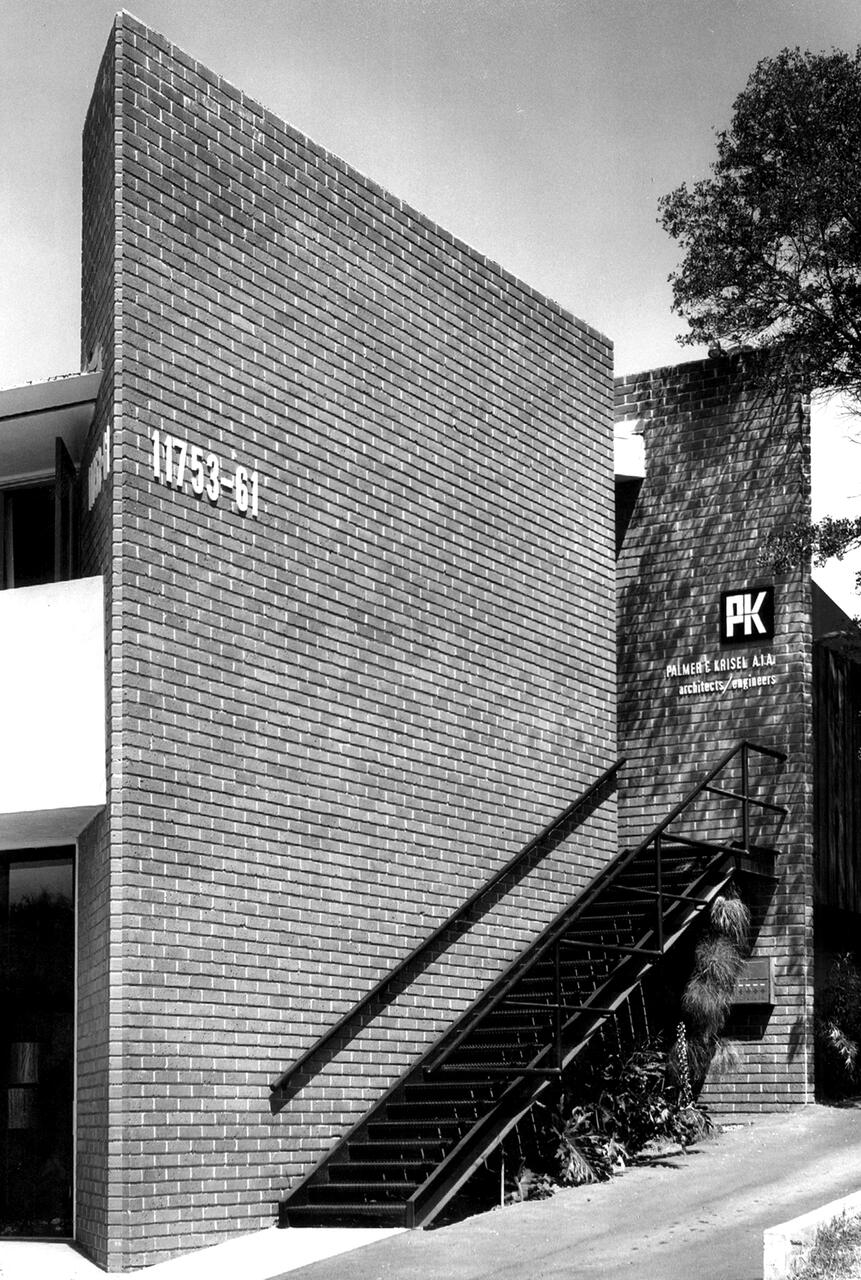 Brick building with a slanted wall featuring numbers "11753-61." A metal staircase leads up to a second section of the building displaying "PK" and "architects/engineers." A tree partially visible on the right. Black and white photograph.