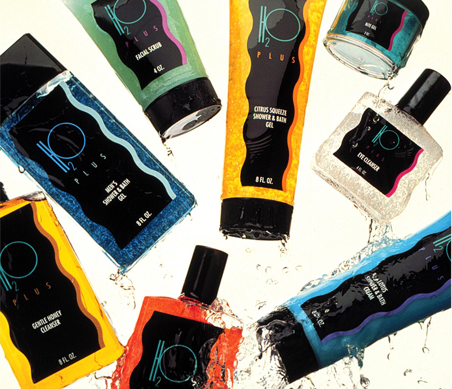 Various H2O Plus skincare products, including shower gels, cleansers, and facial scrub tubes, are creatively arranged on a wet surface, with water splashing around them.