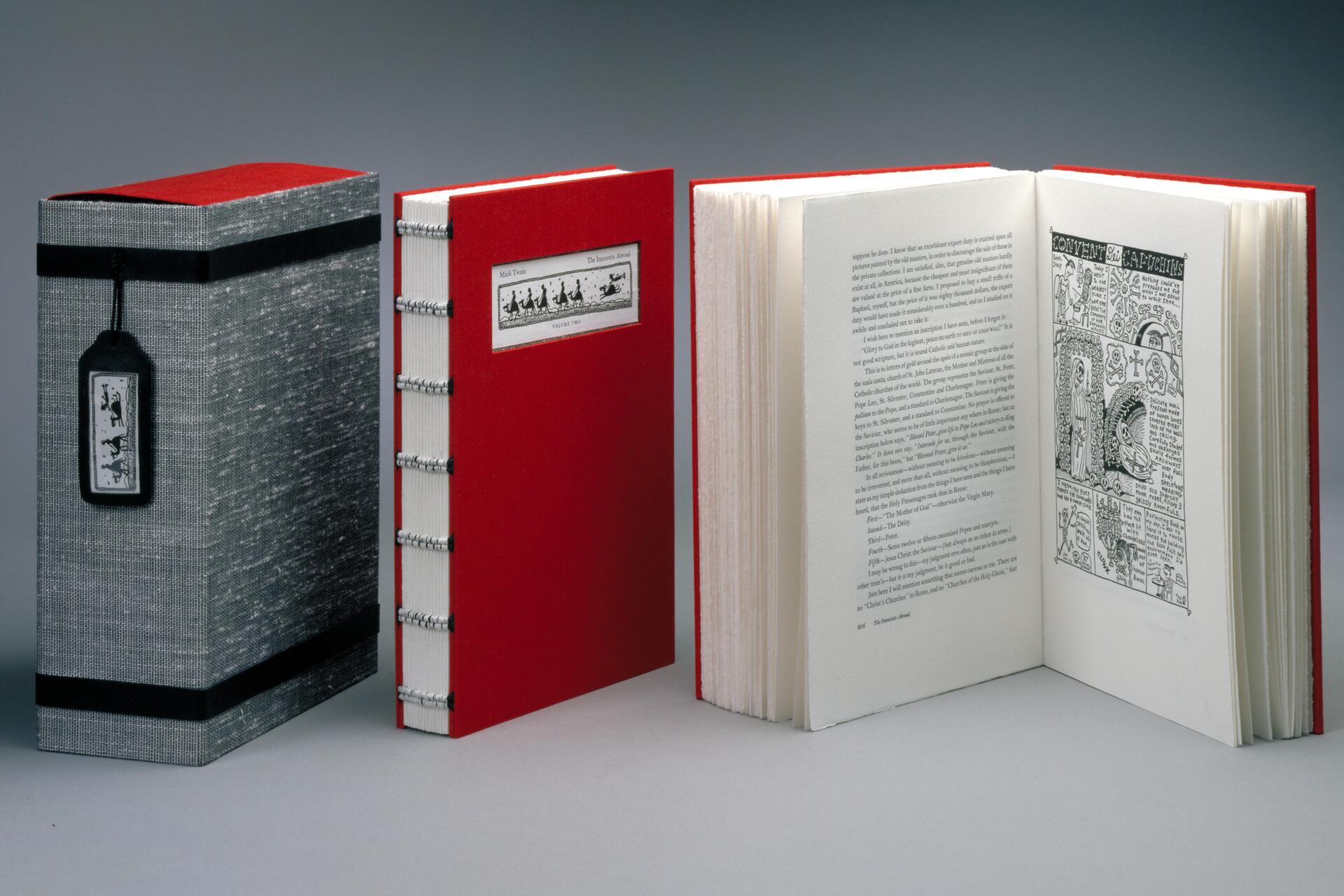A red hardcover book with exposed binding is open to a page featuring text and an illustration. Next to it is the closed book's matching slipcase in gray with a black and white label. The slipcase has a black string with a decorative tag.
