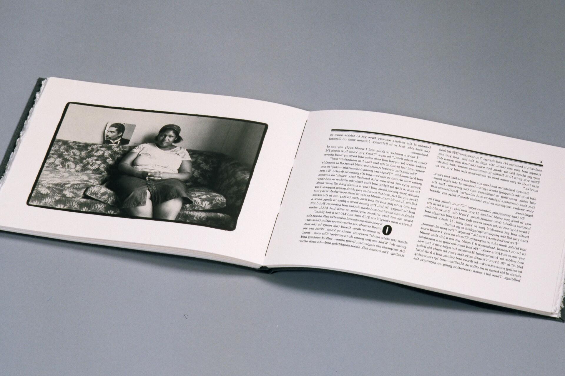 Open book on a gray surface, featuring a black and white photograph of a woman sitting on a patterned couch. On the right page, text is printed.