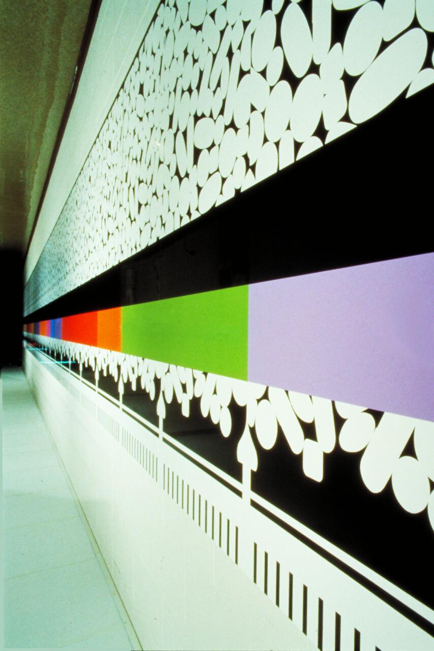 Colorful mural with abstract shapes on a wall. A horizontal black strip divides the vibrant sections, featuring white patterns above and colorful blocks of red, orange, green, and purple below. Geometric designs add a dynamic contrast.