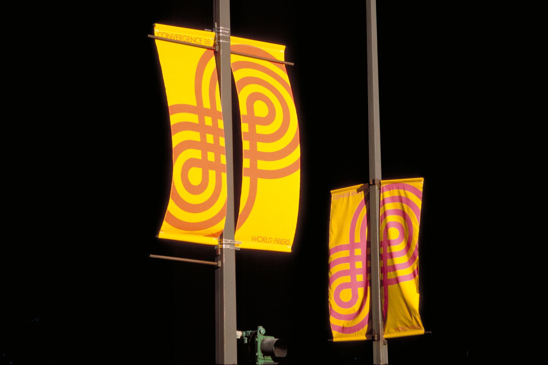 Bright yellow banners with pink geometric patterns are displayed on poles against a dark background.