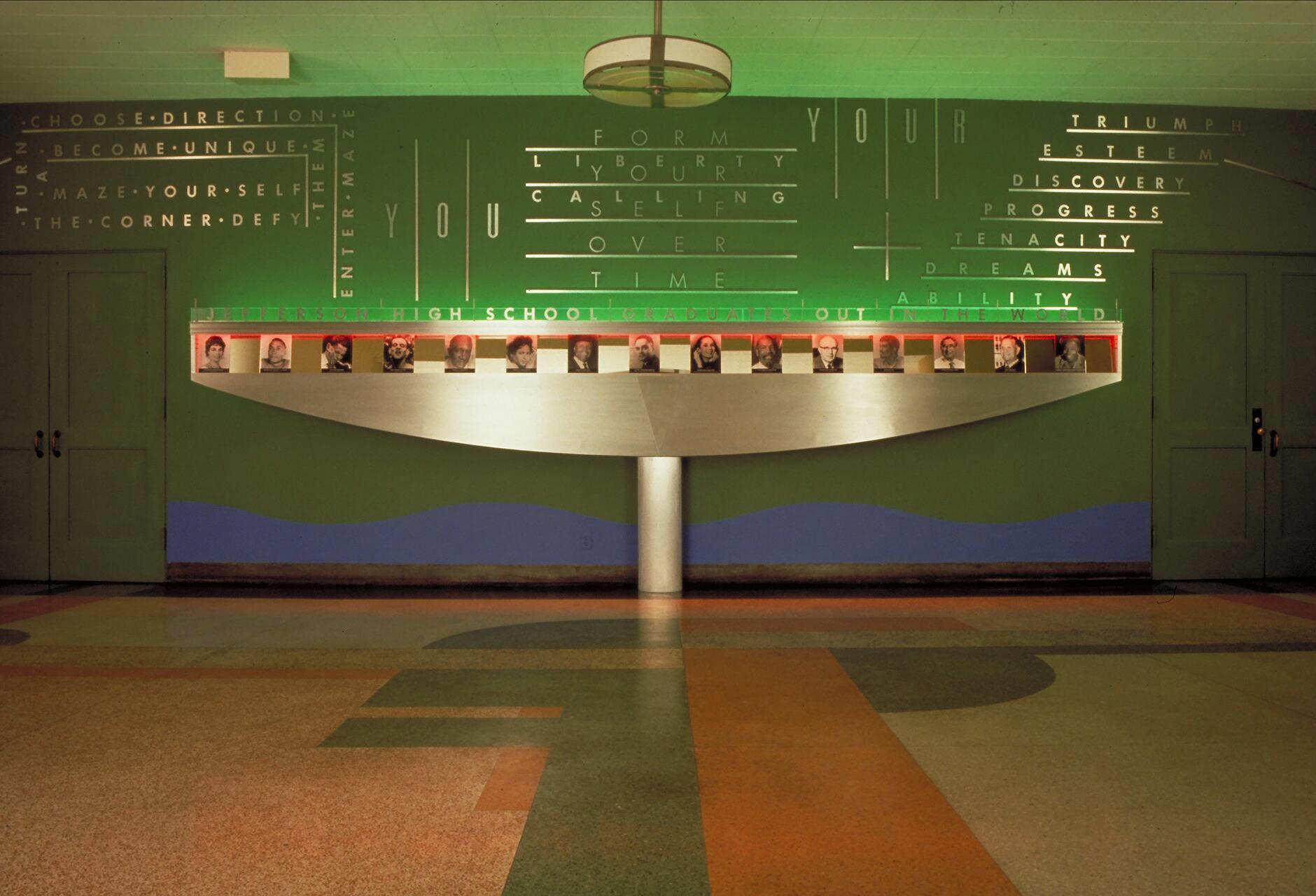 Colorful hallway featuring a green and blue wall with illuminated words and a display with framed photos of various people, flanked by two closed doors. The floor has a geometric pattern in orange, green, and tan.