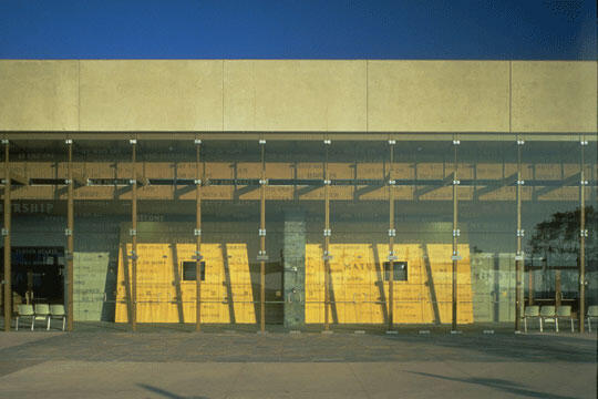 Glass facade of a modern building with visible interior wooden panels and seating. The sun casts strong shadows on the ground, creating a pattern of light and shade.