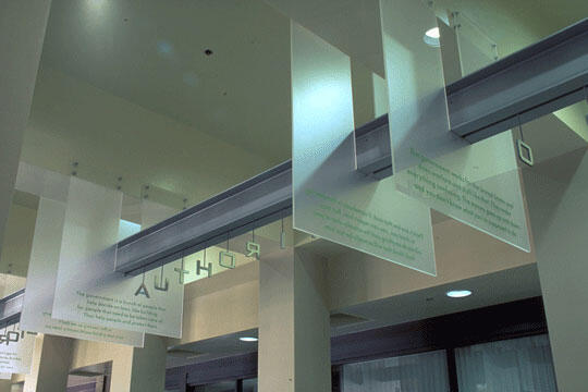 Hanging transparent panels with text, suspended from the ceiling in a modern, well-lit interior. Beige walls with beams crossing overhead, and light shining through windows onto the panels.