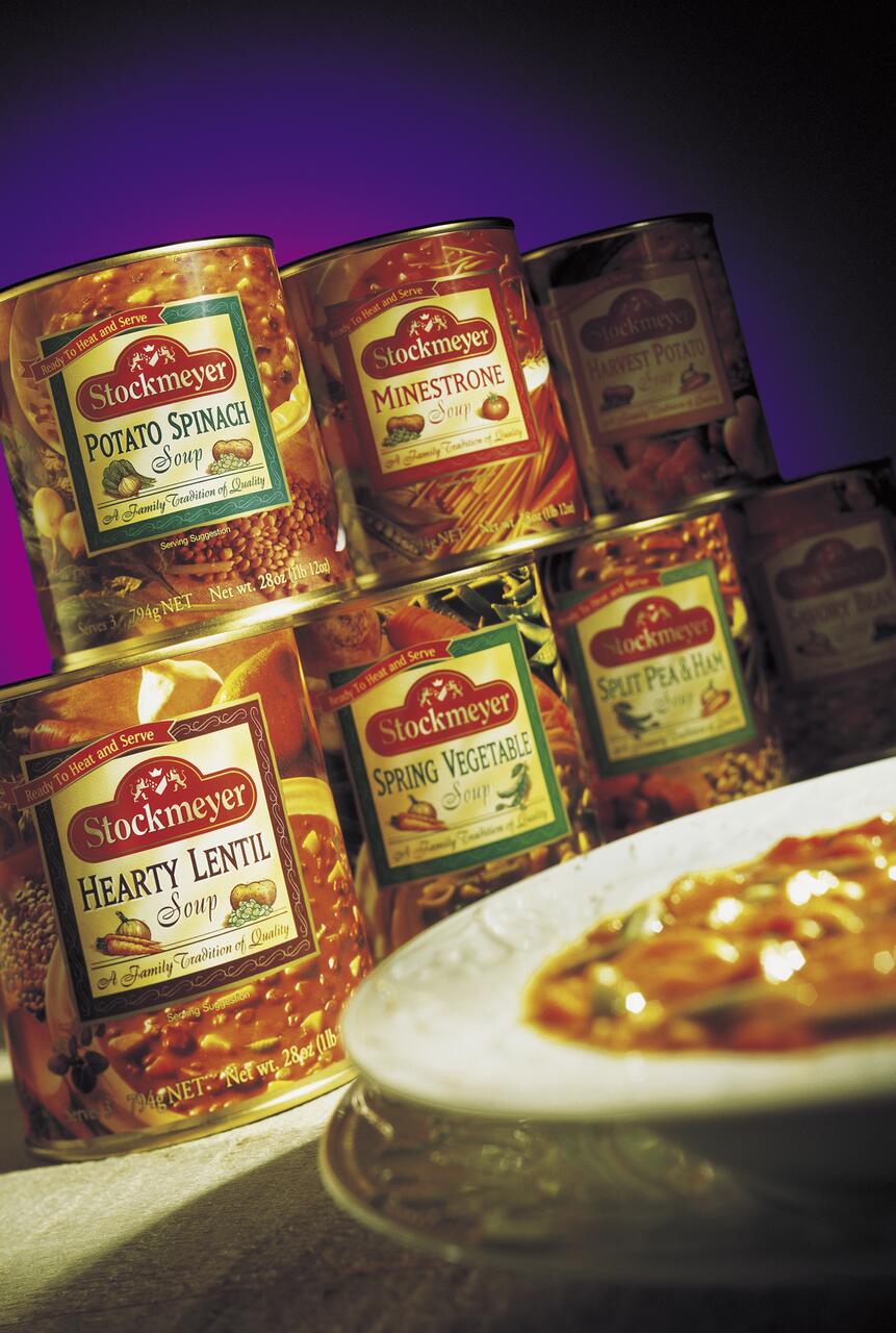 Cans of Stockmeyer soups, including Potato Spinach, Minestrone, and Hearty Lentil, stacked on a surface with vibrant lighting. A bowl of soup is partially visible in the foreground.